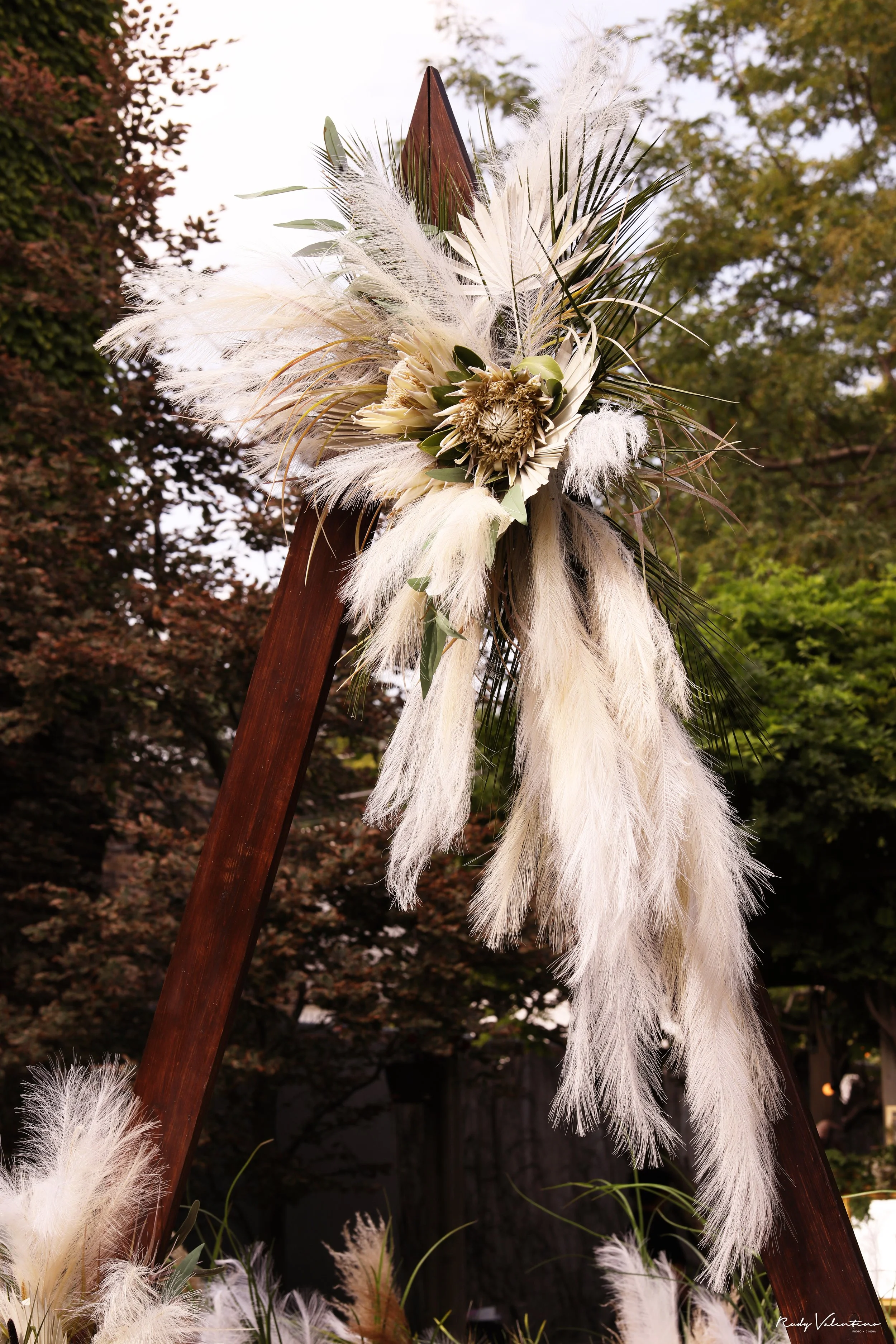 A decorative arrangement of dried flowers, feathers, and greenery attached to a wooden structure outdoors, with trees in the background.