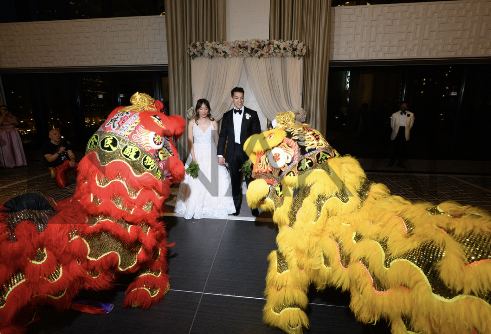 A wedding ceremony with a bride and groom standing beneath a floral arch, surrounded by traditional Chinese lion dancers, one red and one yellow, in a decorated indoor venue.