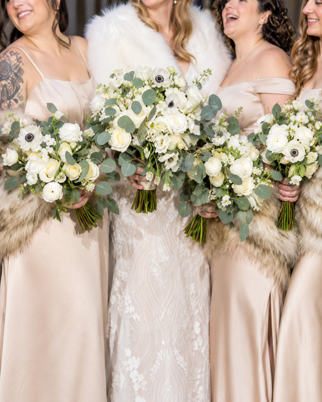 Four women in wedding dresses holding bouquets of white flowers and green foliage, standing together at a wedding.