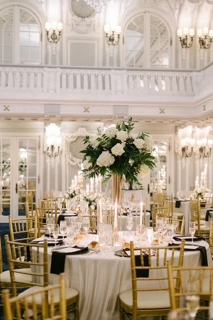 Elegant banquet hall with a tall floral centerpiece of white roses and greenery on a round dining table, surrounded by gold chairs, set with glassware and black napkins, ornate white walls, large windows, and chandeliers.