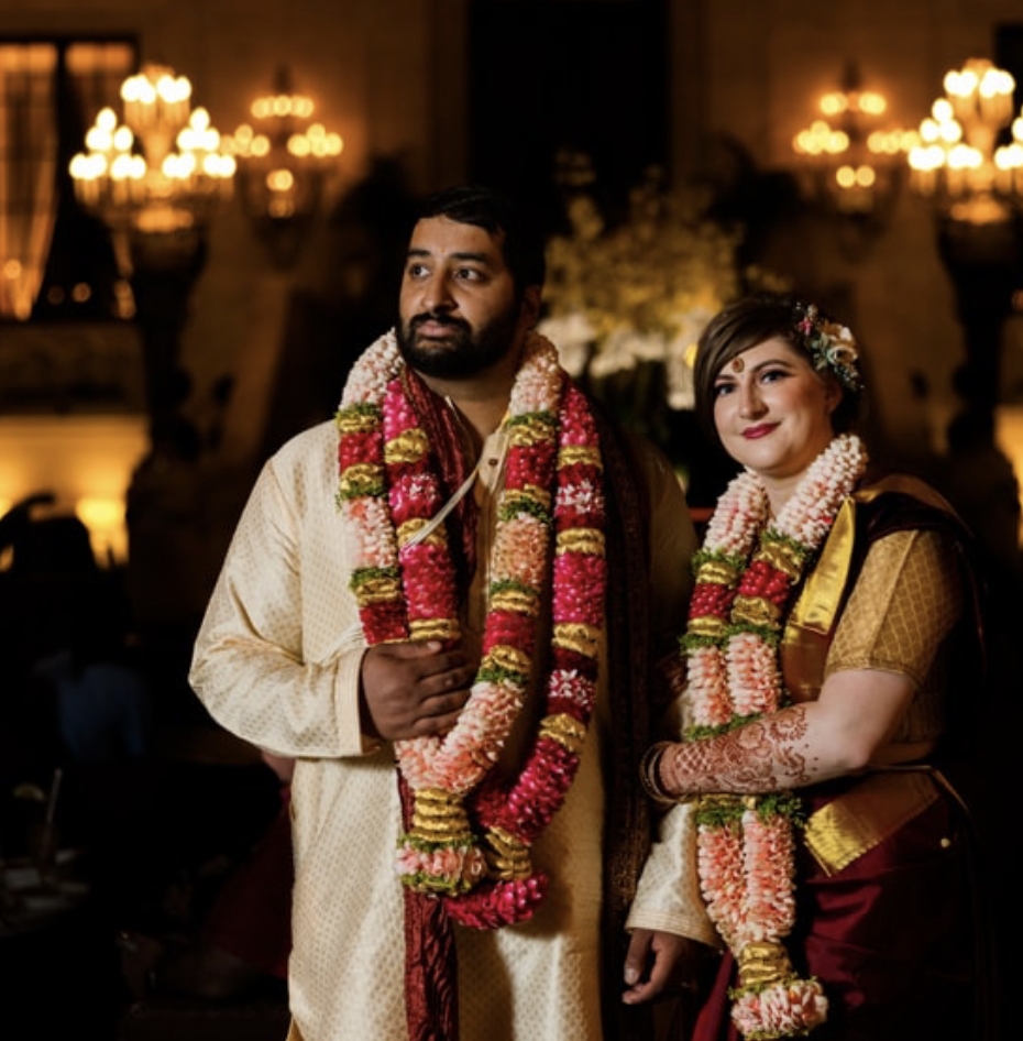 A man and woman dressed in traditional Indian wedding attire, wearing garlands, standing in a decorated venue with warm lighting and chandeliers.