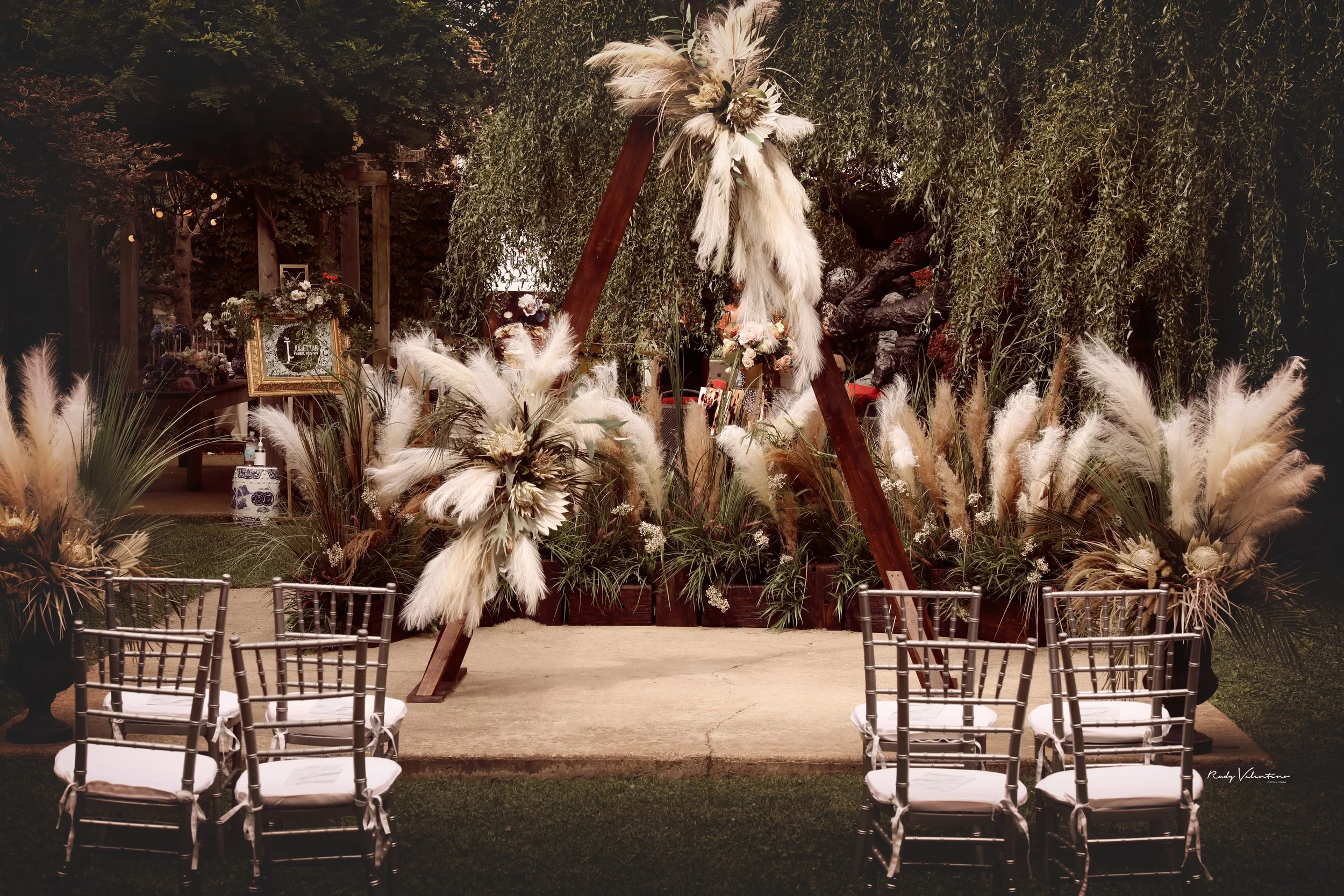 Outdoor wedding ceremony setup with metal chairs, a decorated arch with white and blush flowers, feathers, and greenery, and a lush green backdrop of trees.
