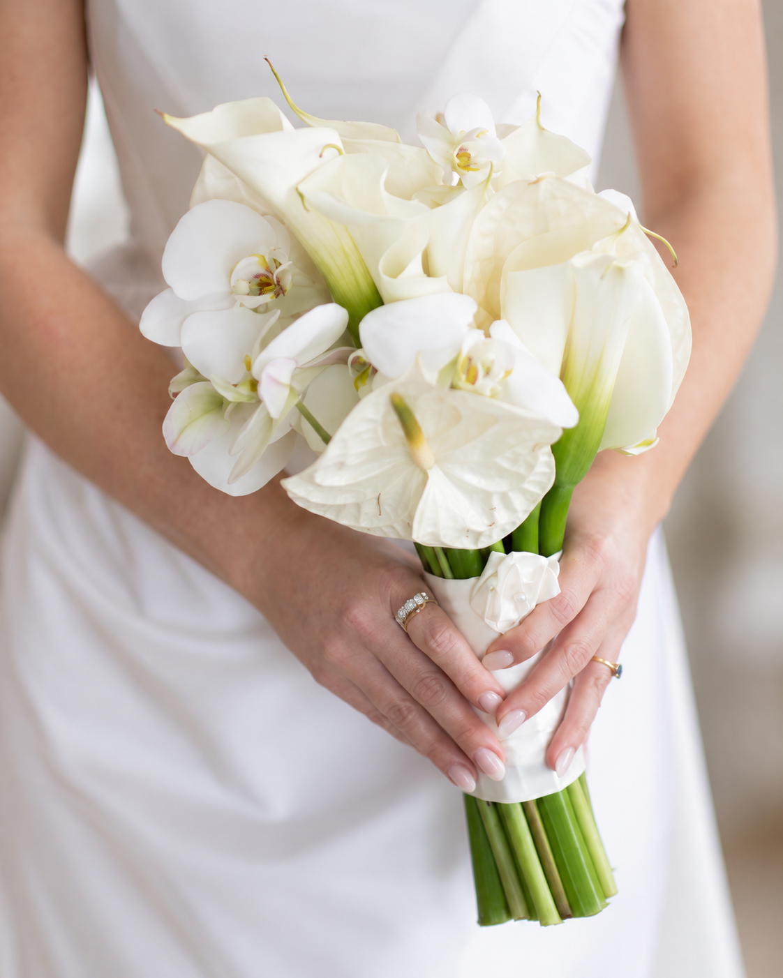 A person in a white dress holding a bouquet of white flowers, including calla lilies and orchids, with their hands clasped around the stems.