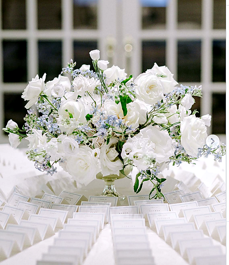 Elegant floral centerpiece with white roses and small blue flowers on a table with place cards.