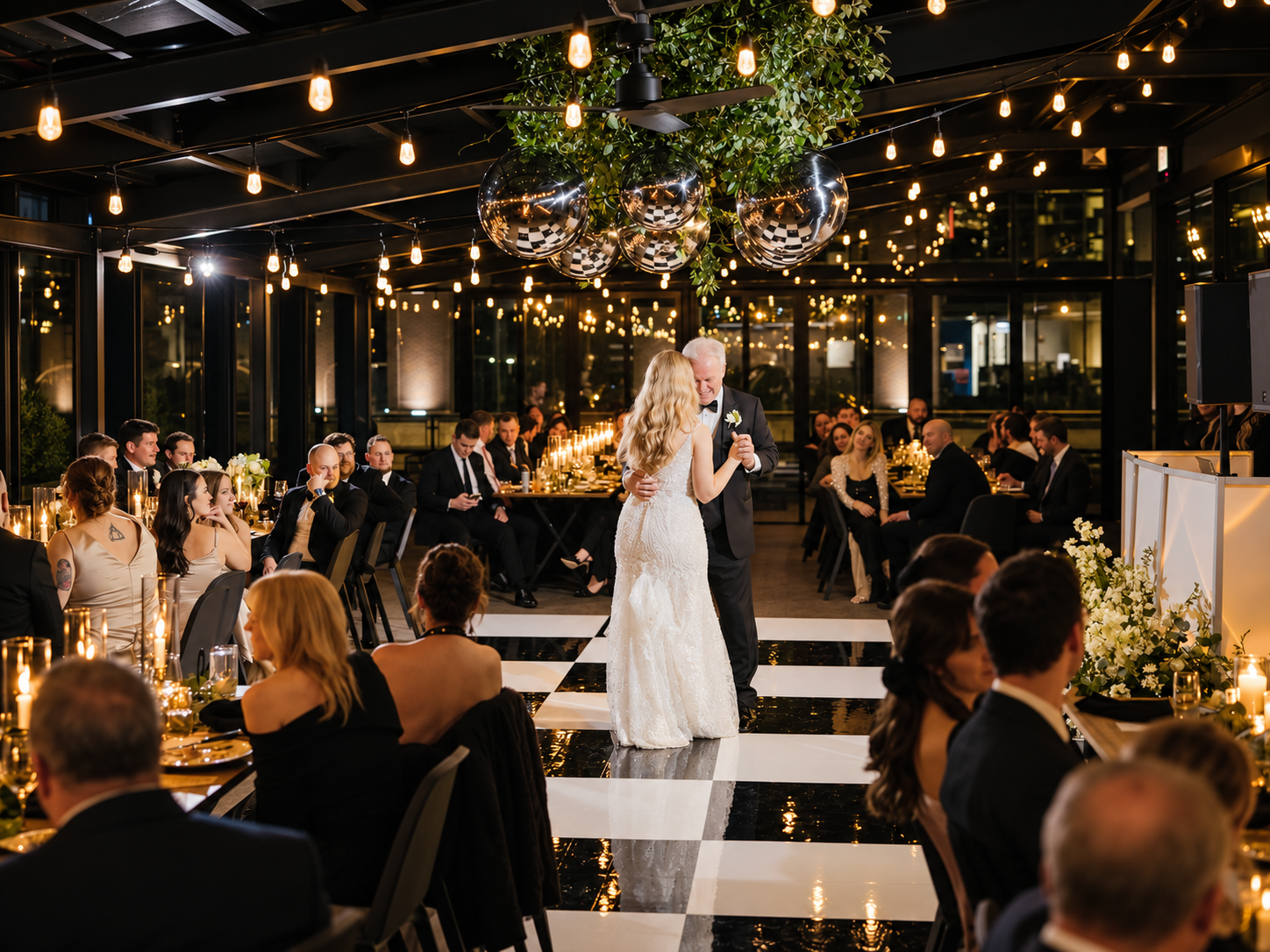 A bride and groom sharing their first dance at a wedding reception, surrounded by seated guests in an elegant, modern venue with black-and-white checkered flooring and warm string lighting.