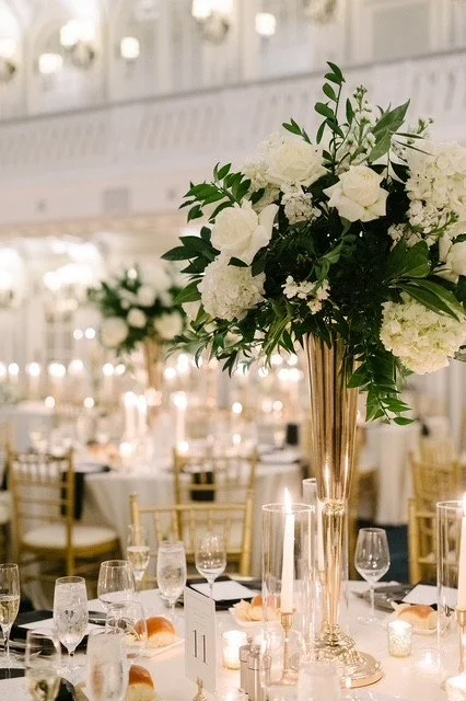 Elegant event table setup with a tall gold vase holding white flowers and greenery, surrounded by wine glasses, water glasses, bread rolls, and candlelight in a decorated banquet hall.