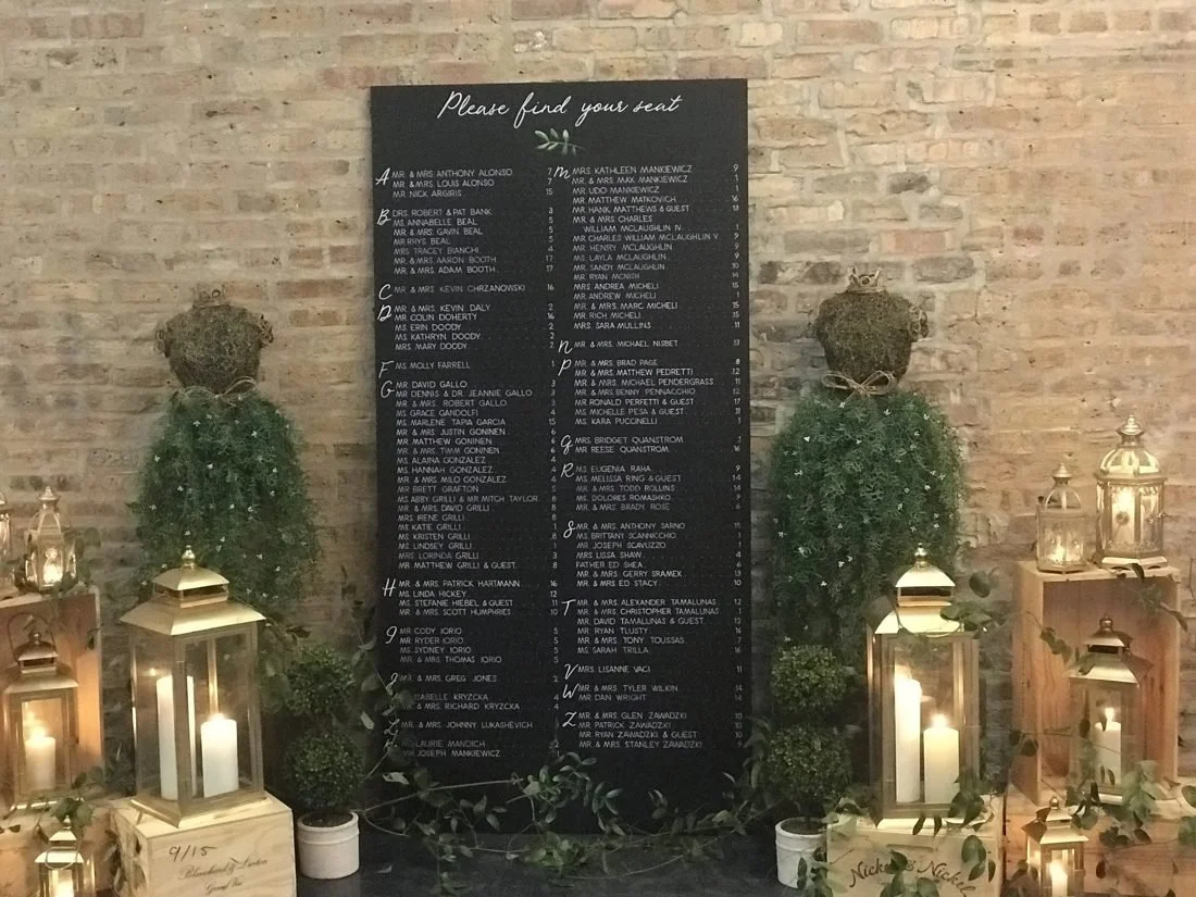 A black wedding seating chart with white cursive text hangs on a brick wall, surrounded by greenery and candles in lanterns on wooden crates.