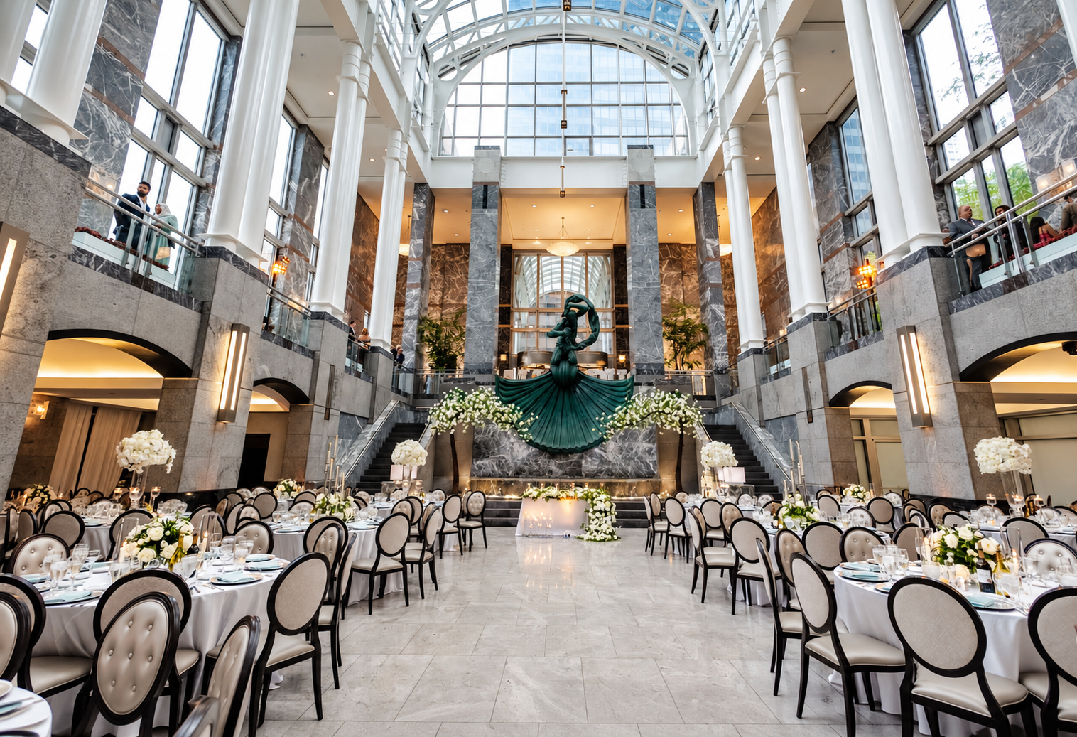 Elegant banquet hall with multiple round tables decorated with white flowers and candles, a grand staircase, and a large abstract sculpture of a woman in a flowing dress at the center, with tall windows and marble walls.