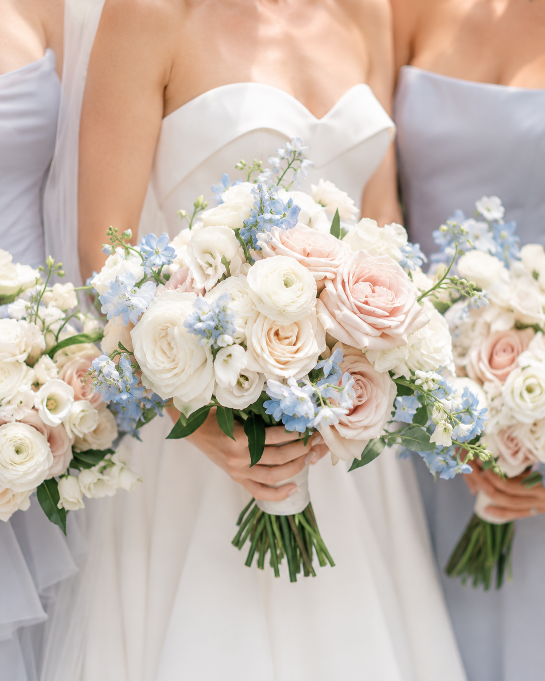 A woman in a white strapless wedding dress holding a bouquet of white, cream, and blush roses with blue accent flowers and green leaves.
