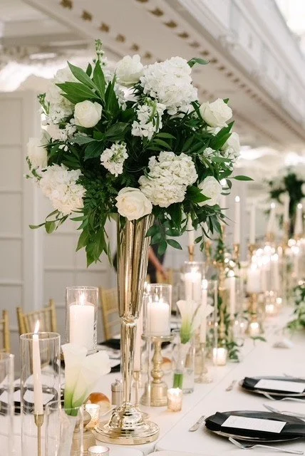 Elegant floral centerpiece with white roses and hydrangeas on a wedding reception table, surrounded by candles and table setting.