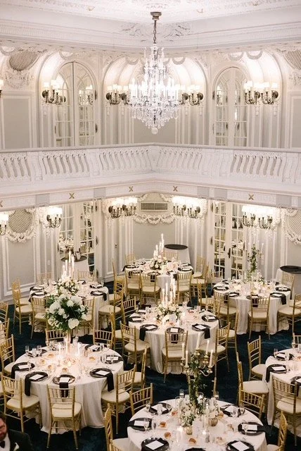 Elegant banquet hall decorated for a formal event with round tables, white tablecloths, floral centerpieces, and gold chairs. The room features a chandelier, wall sconces, tall windows, and ornate crown molding.