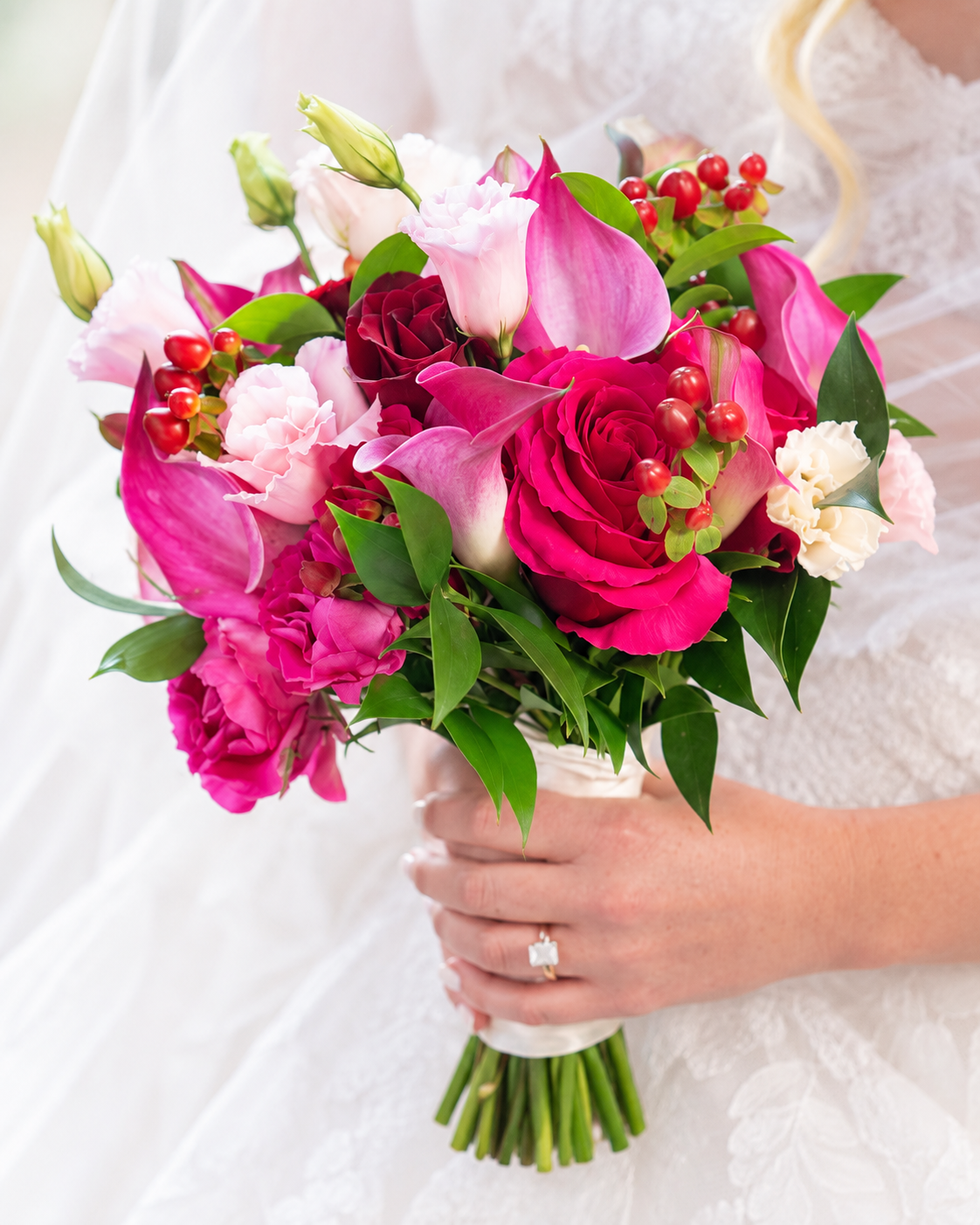 A bride in a white dress holding a colorful bouquet of pink and red roses, calla lilies, and greenery.