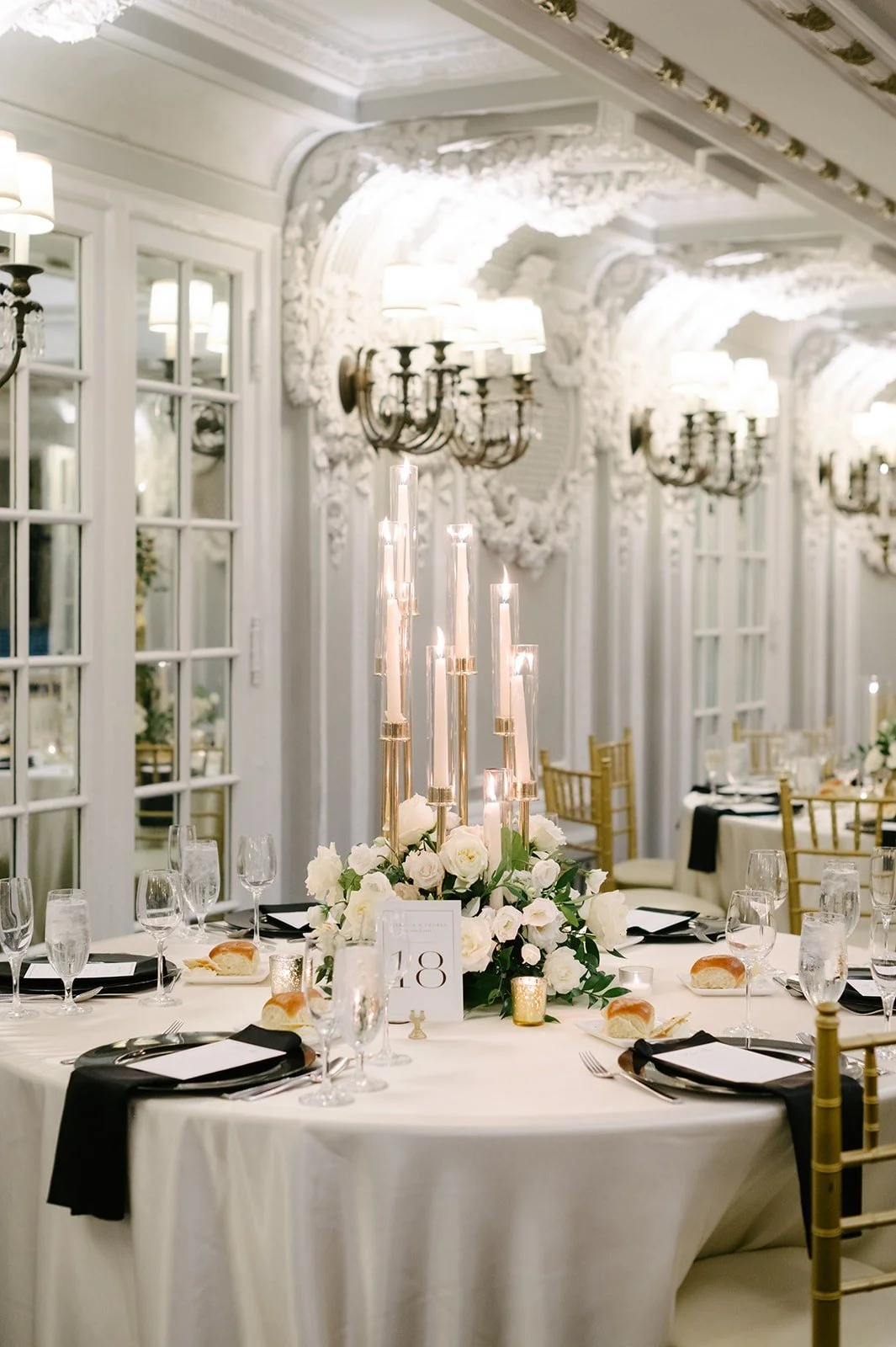 Elegant banquet hall decorated with tall white candles, white and cream roses, and gold accents. Well-set tables with black napkins, glassware, and bread rolls, surrounded by gold chairs. The room features ornate white woodwork, wall-mounted light fi