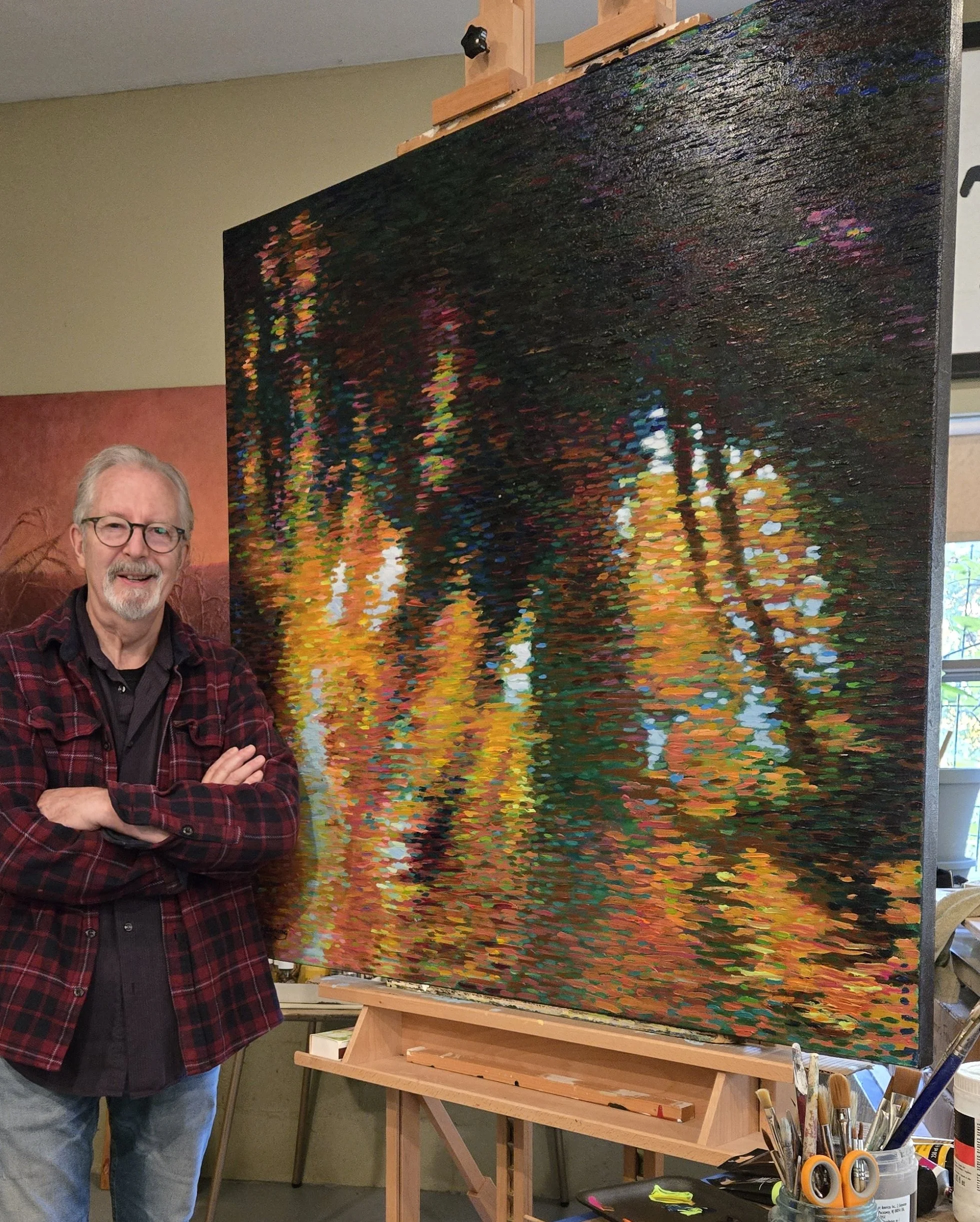 Artist Stephen Henning standing in his studio beside a large abstract landscape painting titled Jubilation, featuring luminous reflections of gold, green, and blue.