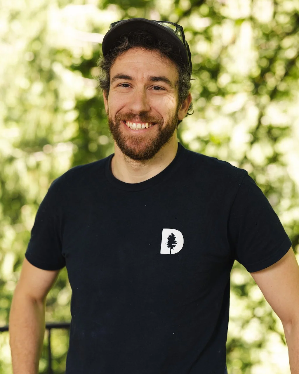 Smiling person wearing a black t-shirt with a tree logo, standing outdoors with a blurred green background.