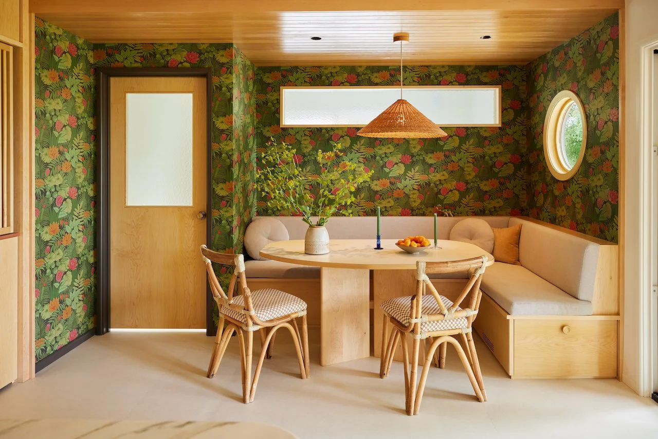 Cozy dining nook with floral wallpaper, round wooden table, corner bench seating, rattan chairs, pendant light, and decorative plants.