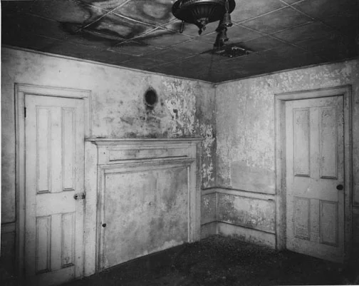 This 1962 photo shows the hole over the fireplace for a potbelly stove at the rear parlor, a later addition that was more efficient than the fireplaces for heating. The stoves were removed from the room each summer and stored.
