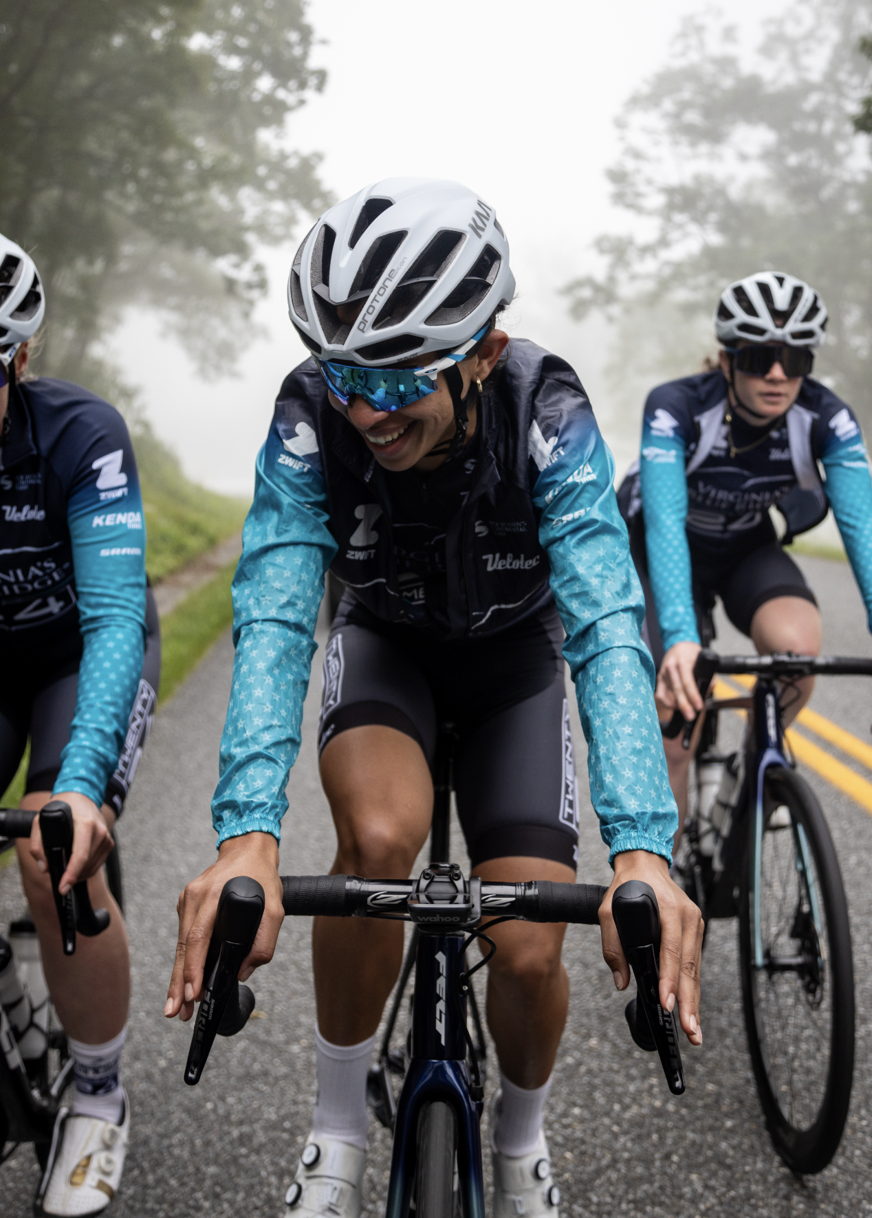 Three female cyclists riding on a foggy road surrounded by trees, wearing cycling helmets, sunglasses, and matching cycling jerseys.