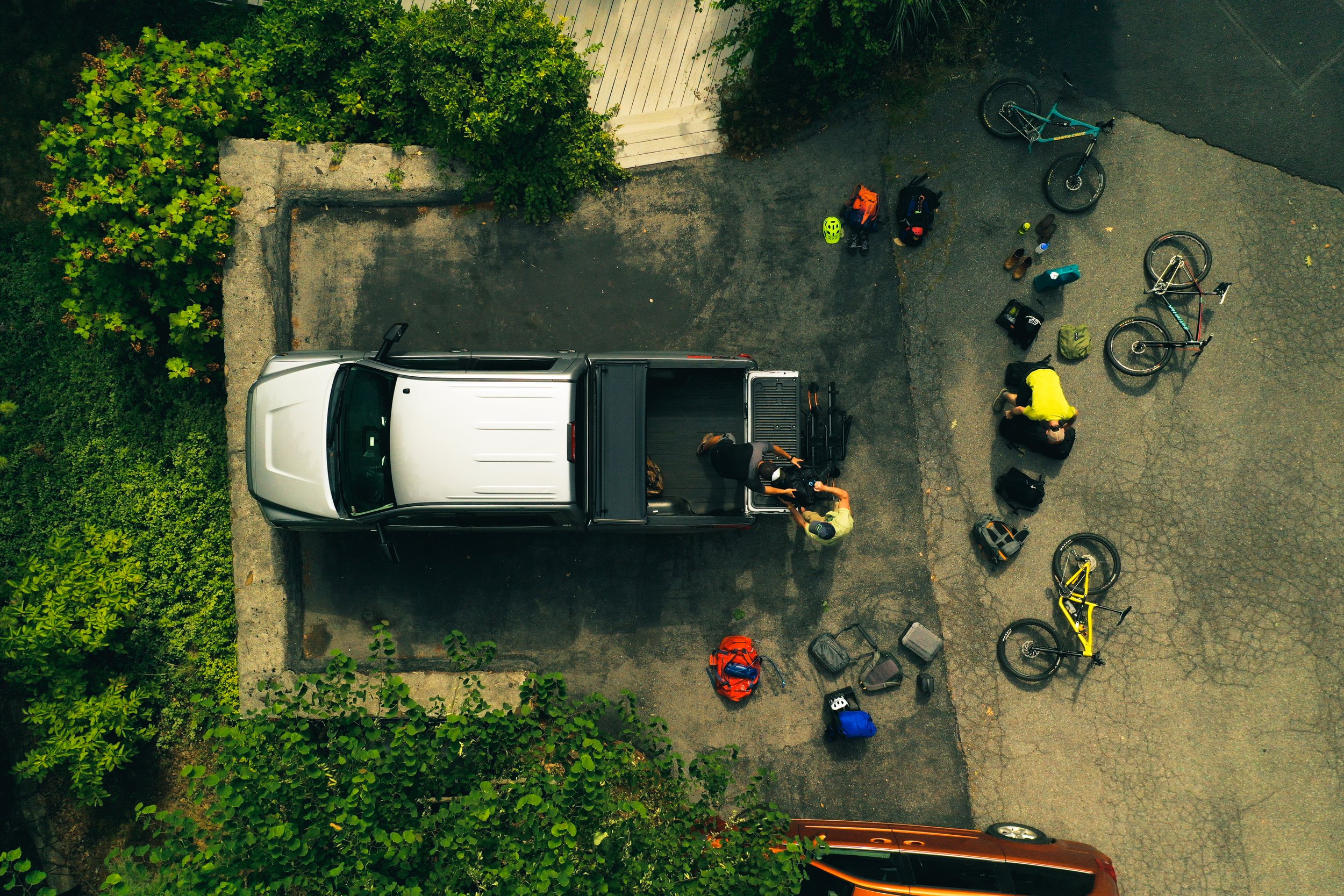 Aerial view of a driveway with a white pickup truck, bicycles, helmets, backpacks, and equipment scattered around. Two people are near the truck, one in a yellow shirt. Green bushes border the driveway.
