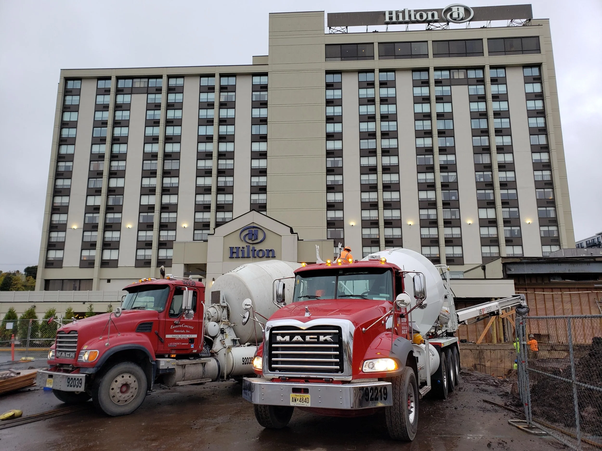 Construction site in front of a Hilton hotel, with two red cement trucks, construction workers, and machinery on muddy ground.
