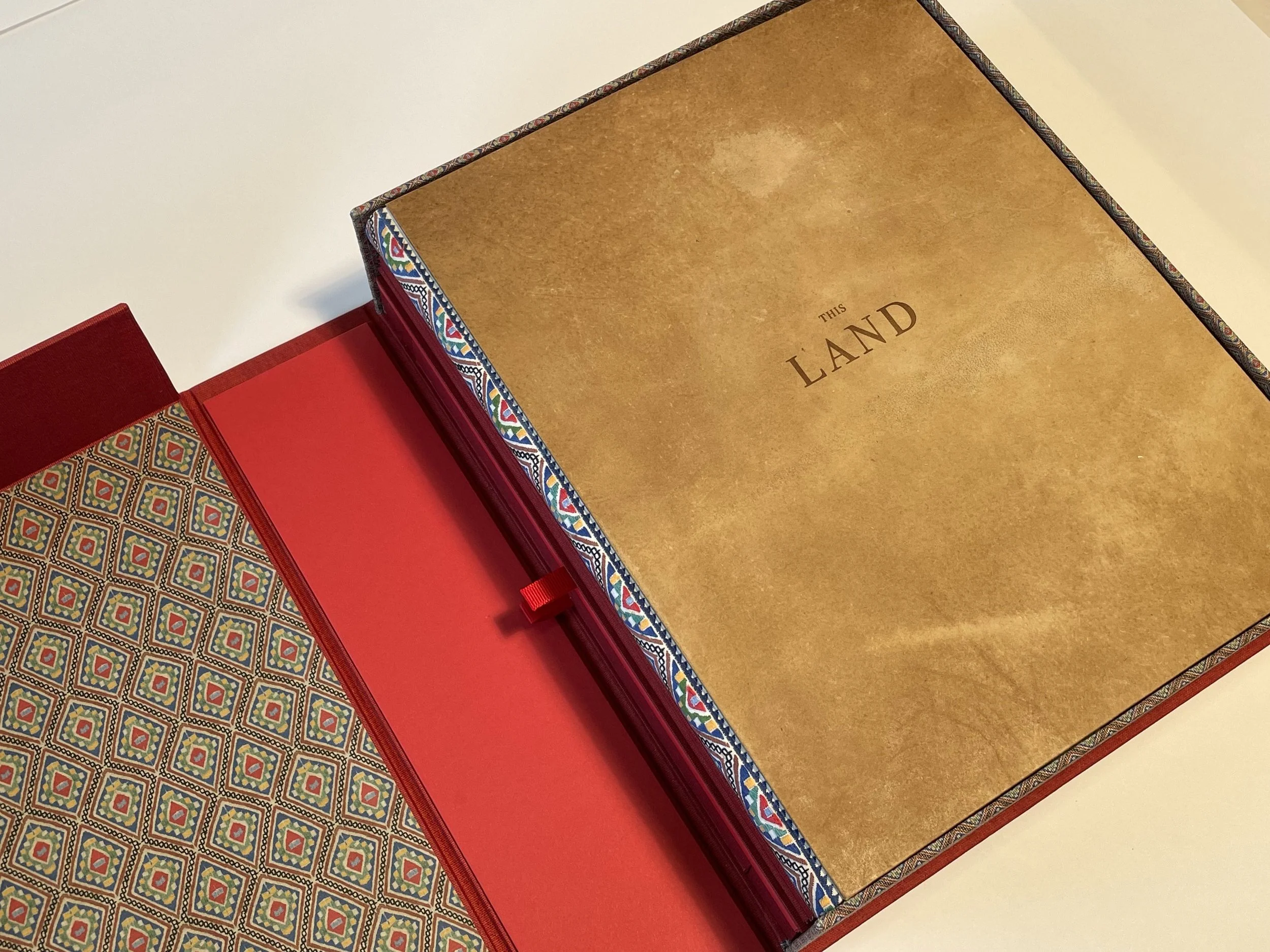 Open box with colorful intricate pattern on outer border, and 'This Land' written on inside lid, with red interior and background.