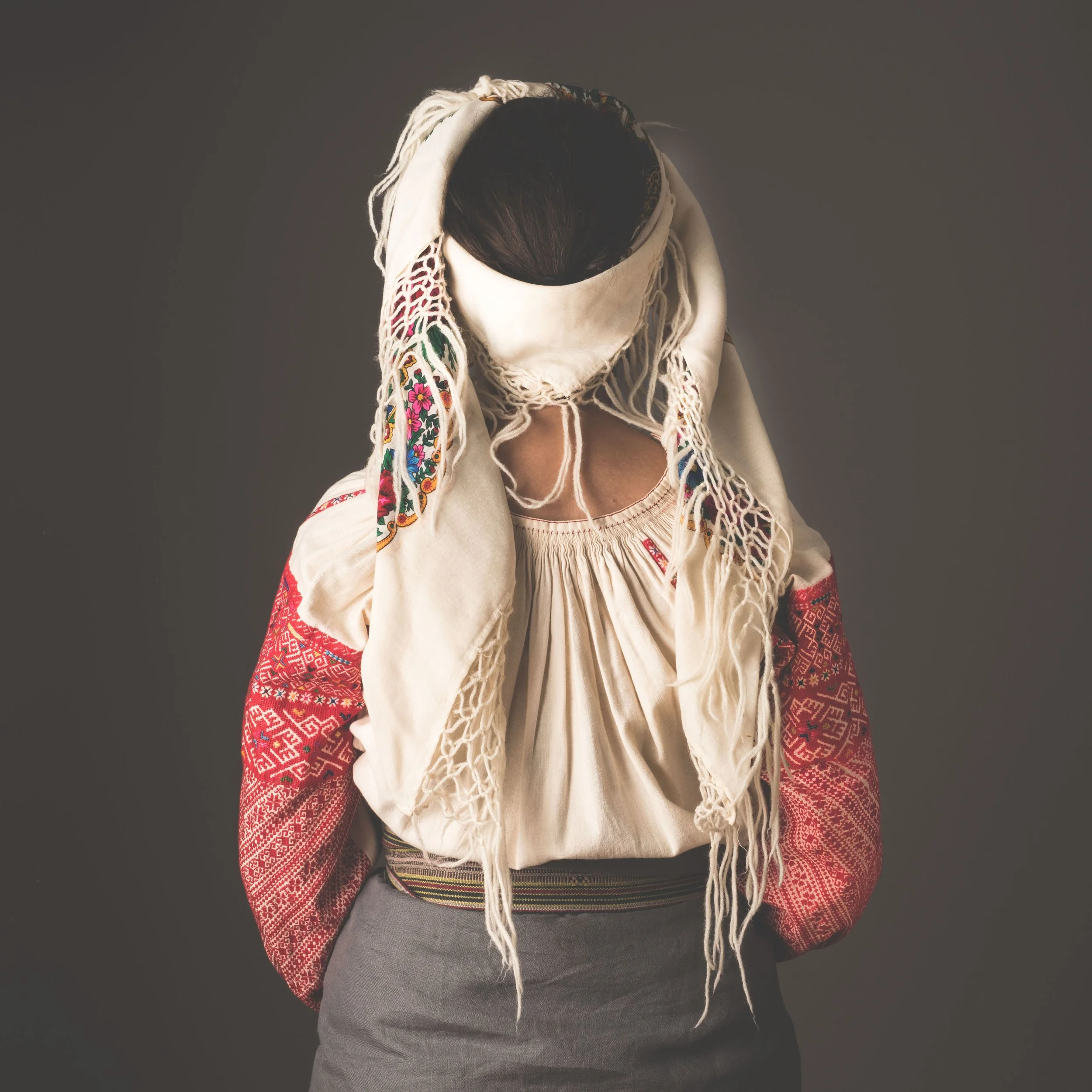 A woman wearing traditional embroidered clothing with a headscarf covering her face, standing against a plain background.