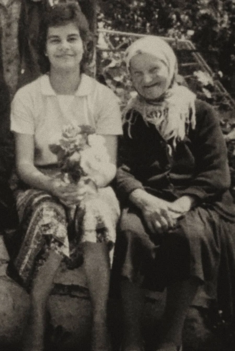 Two women sitting outdoors, one with dark, curly hair in a white top and patterned pants, and the other with light hair in a dark jacket and scarf, smiling at the camera.