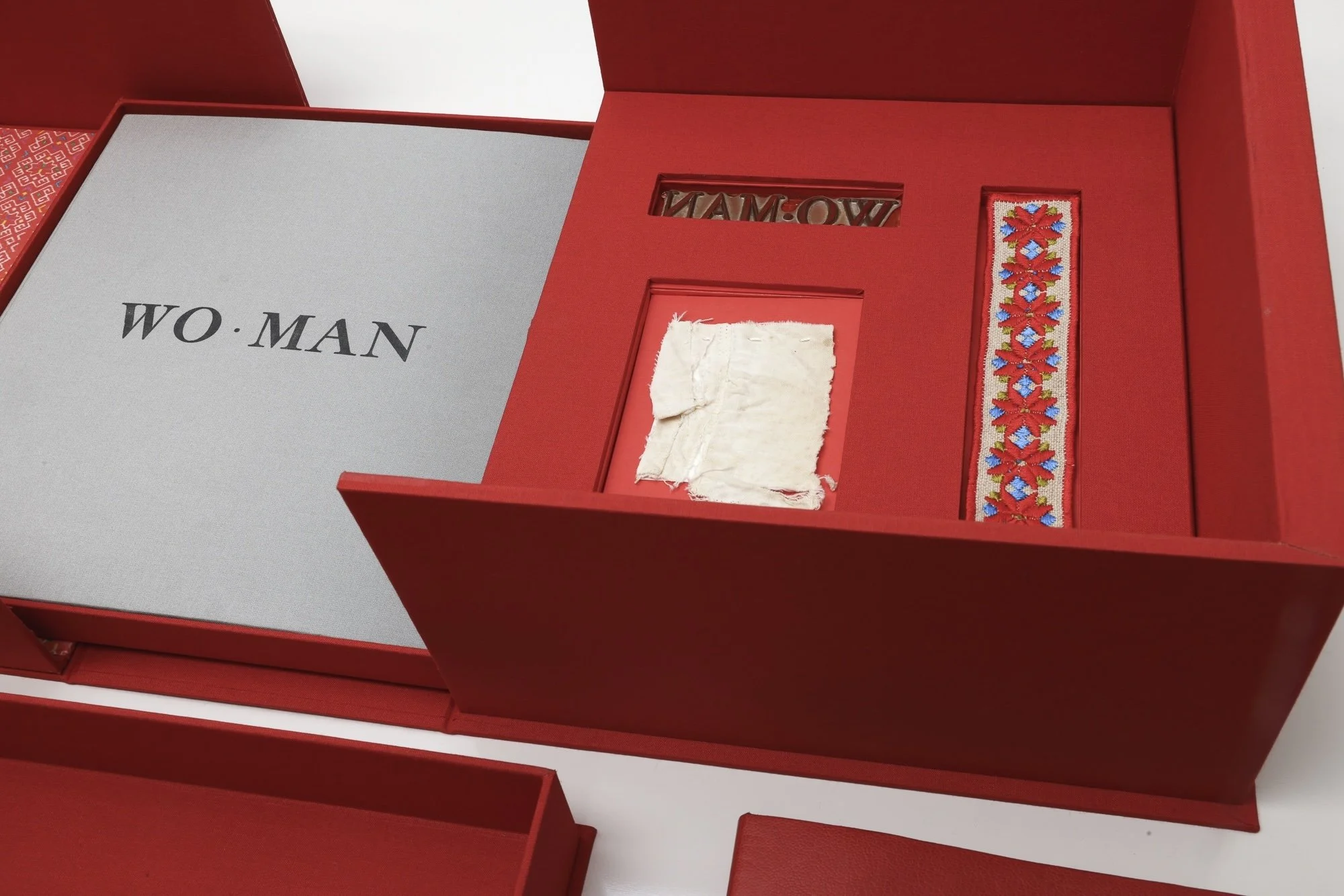 Empty red jewelry box with compartments, one containing a torn piece of white fabric, another a embroidered band with red, blue, and yellow flowers, and a small cutout with the word 'WOW' inside.
