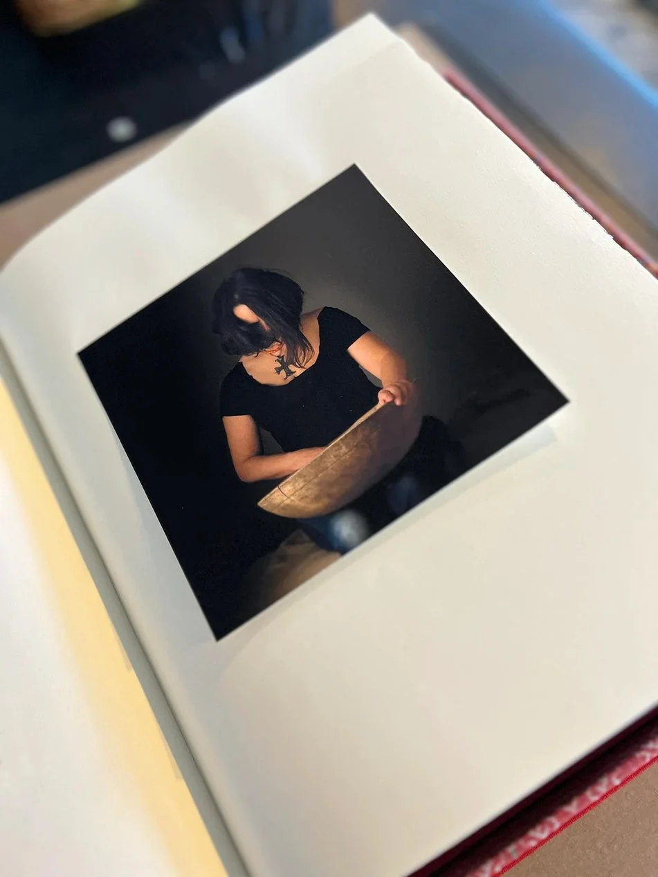 Photograph of a woman with dark hair looking down, wearing a black dress, sitting at a small wooden table inside a dark space.