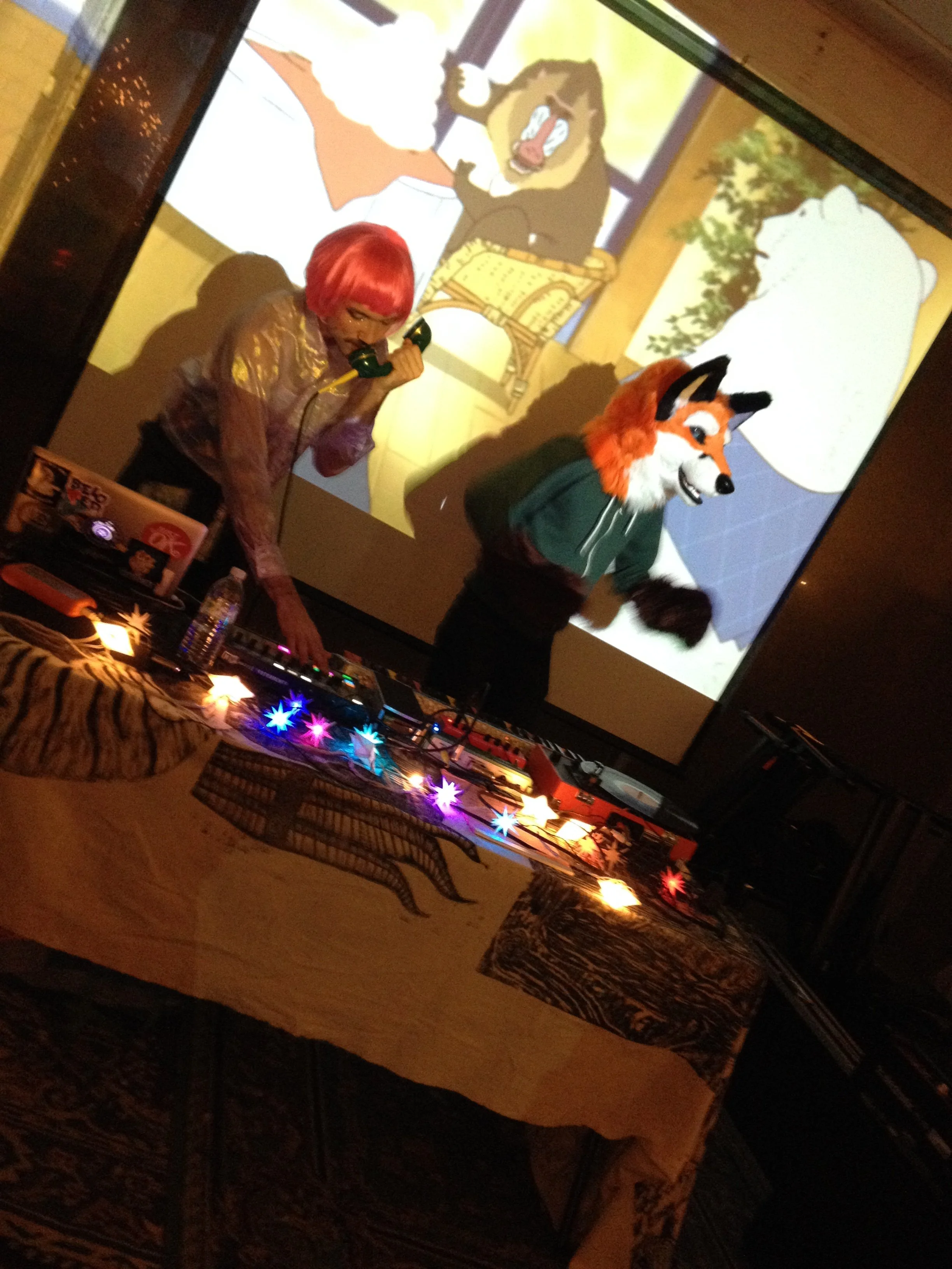 Two people standing behind a table one with a pink wig and one wearing a fursuit fox head DJing in front of a projection screen
