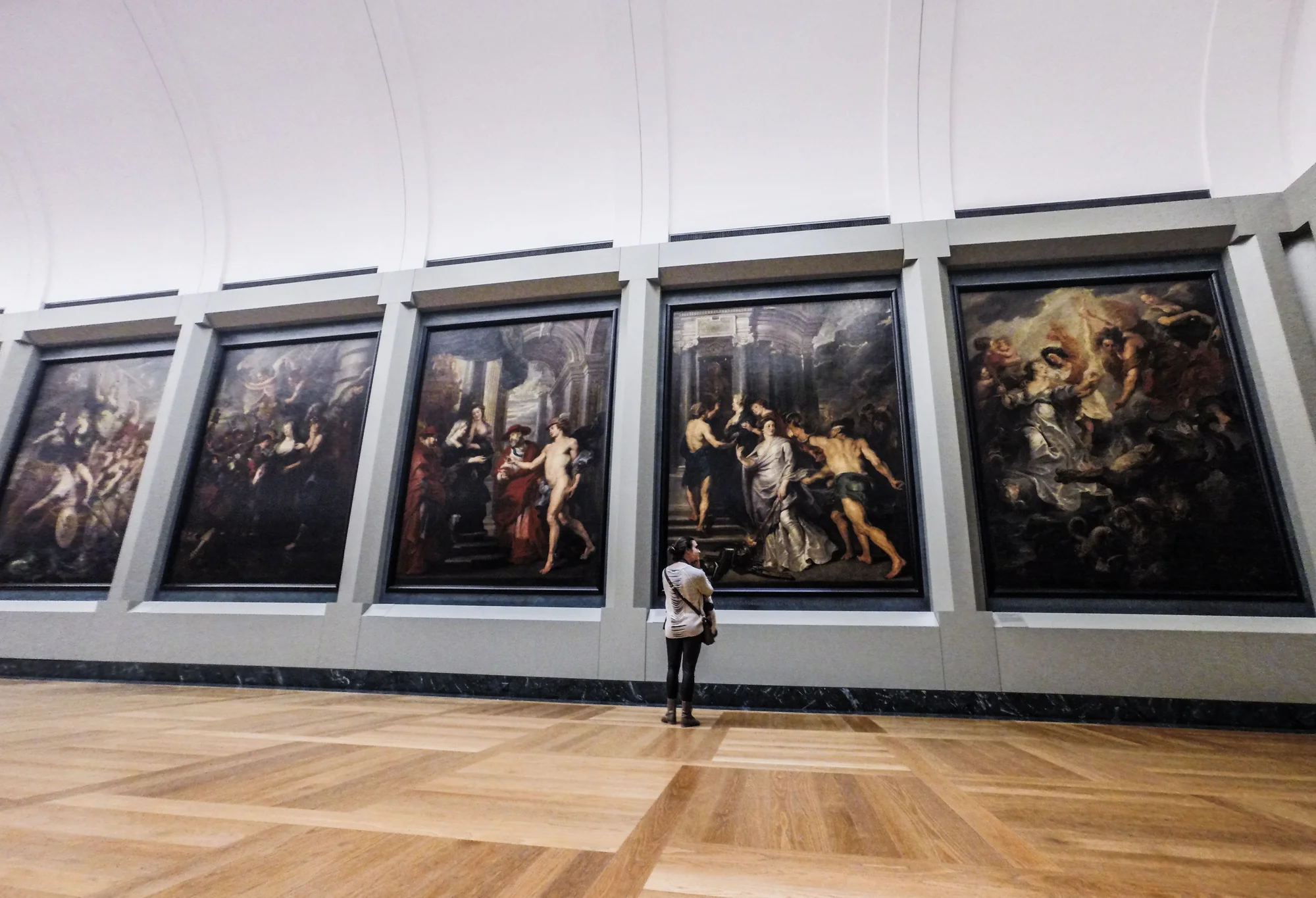 A person stands in front of large classical paintings in an art museum gallery, with a wooden floor and a high, curved ceiling.