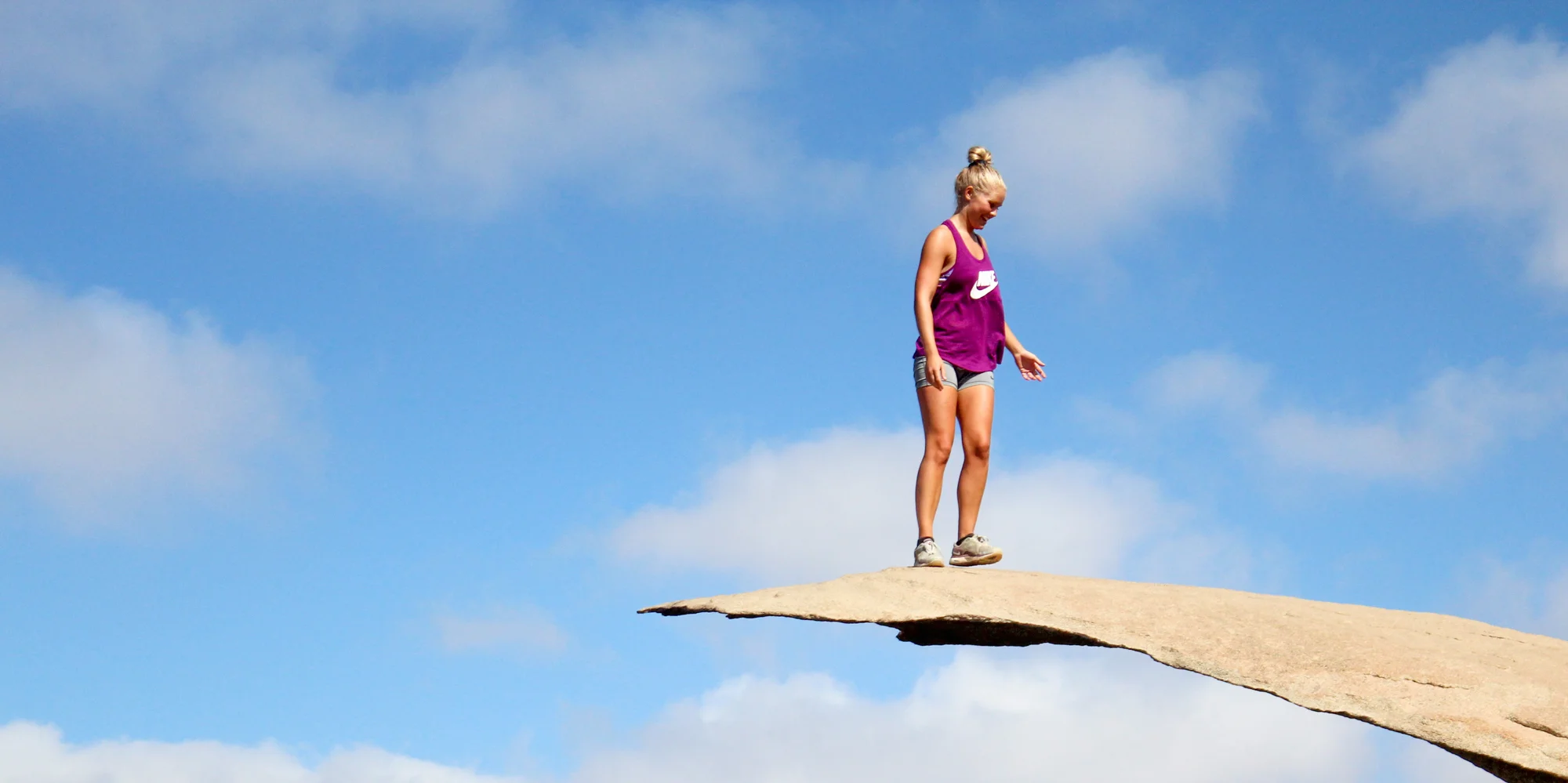 A woman in a purple tank top and shorts walking on a thin rock ledge against a blue sky with clouds.