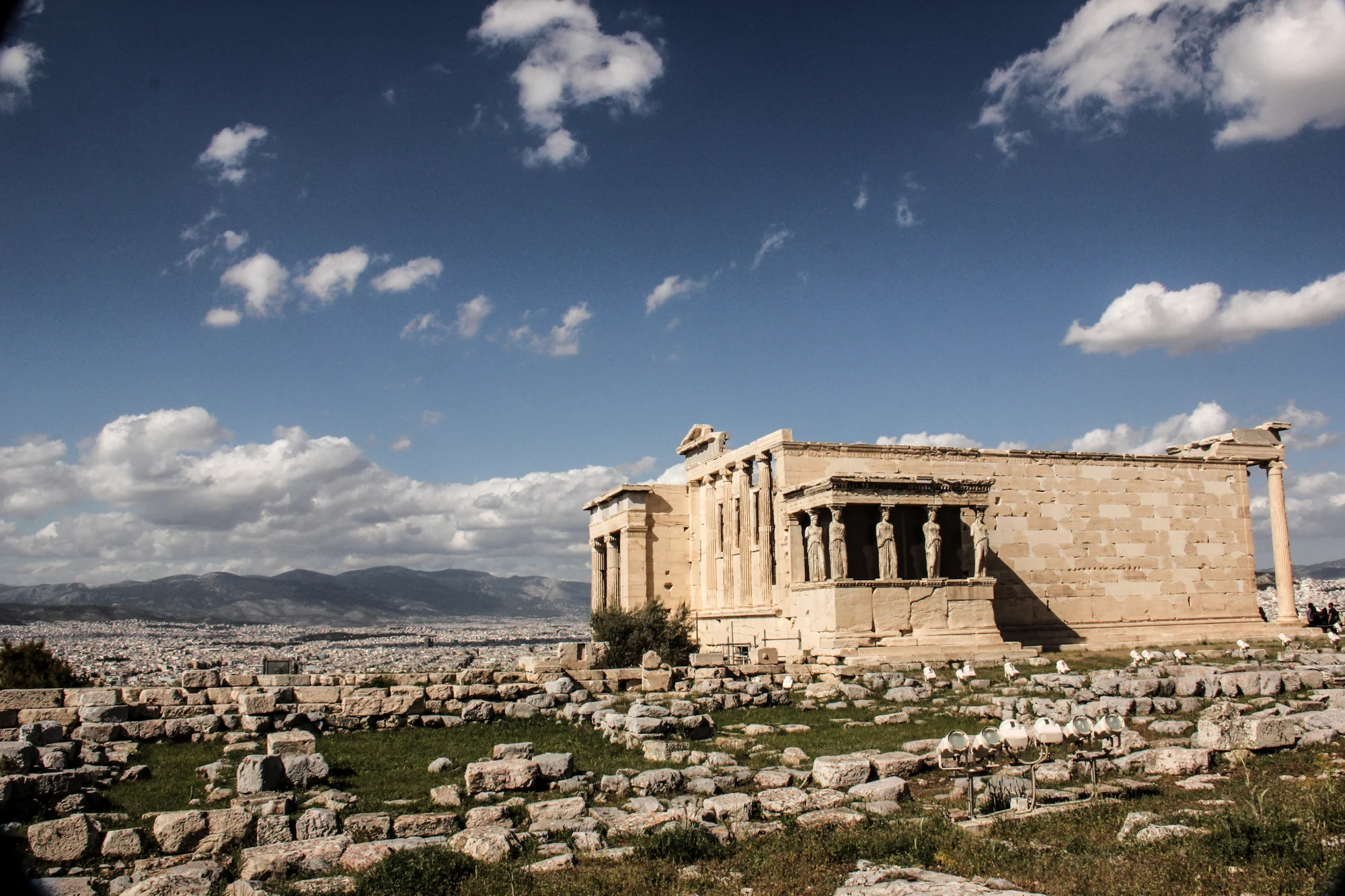 Ancient Greek Parthenon temple on the Acropolis in Athens, Greece, with a blue sky and some clouds in the background, and rocky terrain in the foreground.