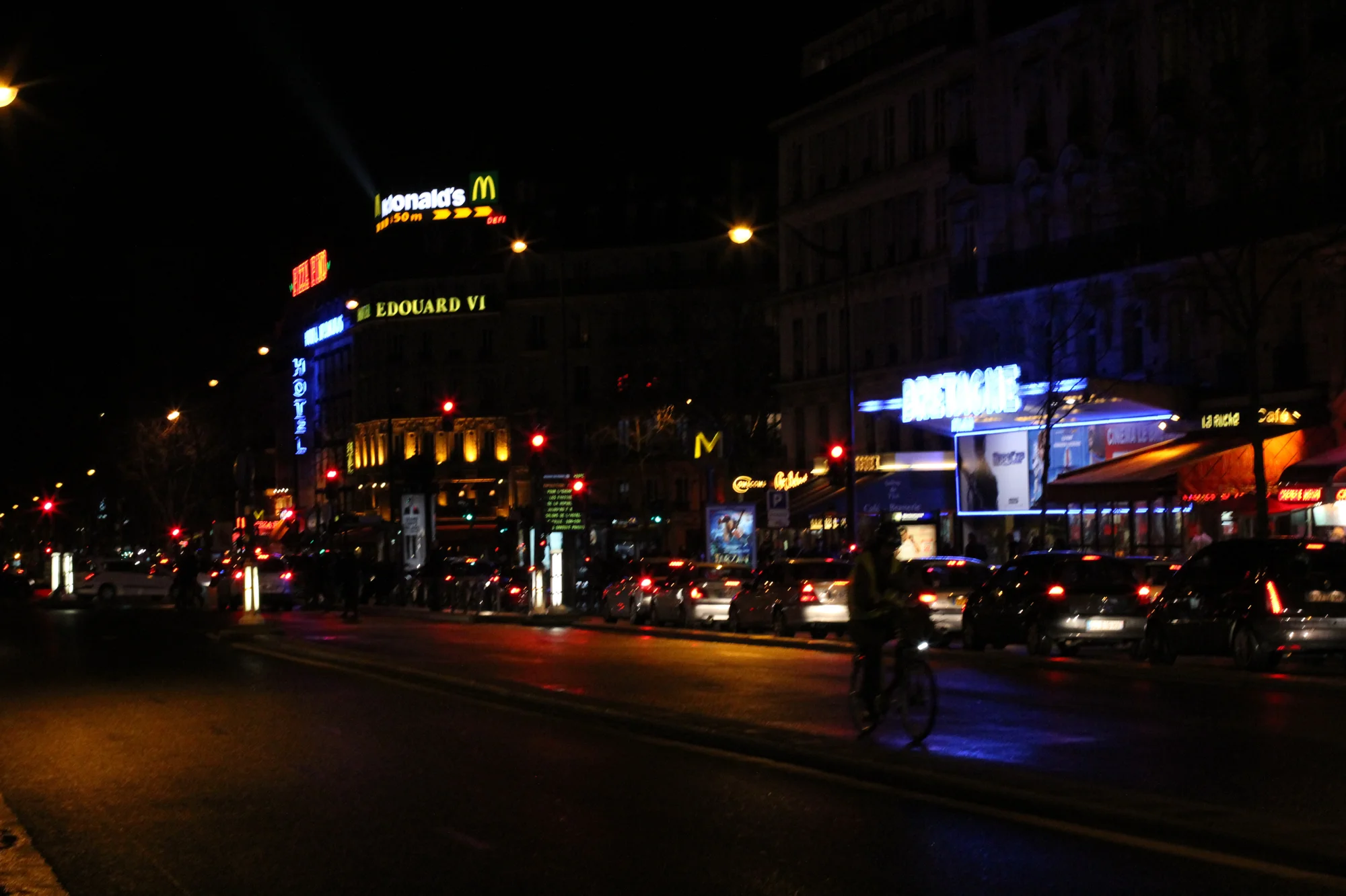 Nighttime city street scene with illuminated signs, cars parked along the street, and a cyclist riding on the bike lane.