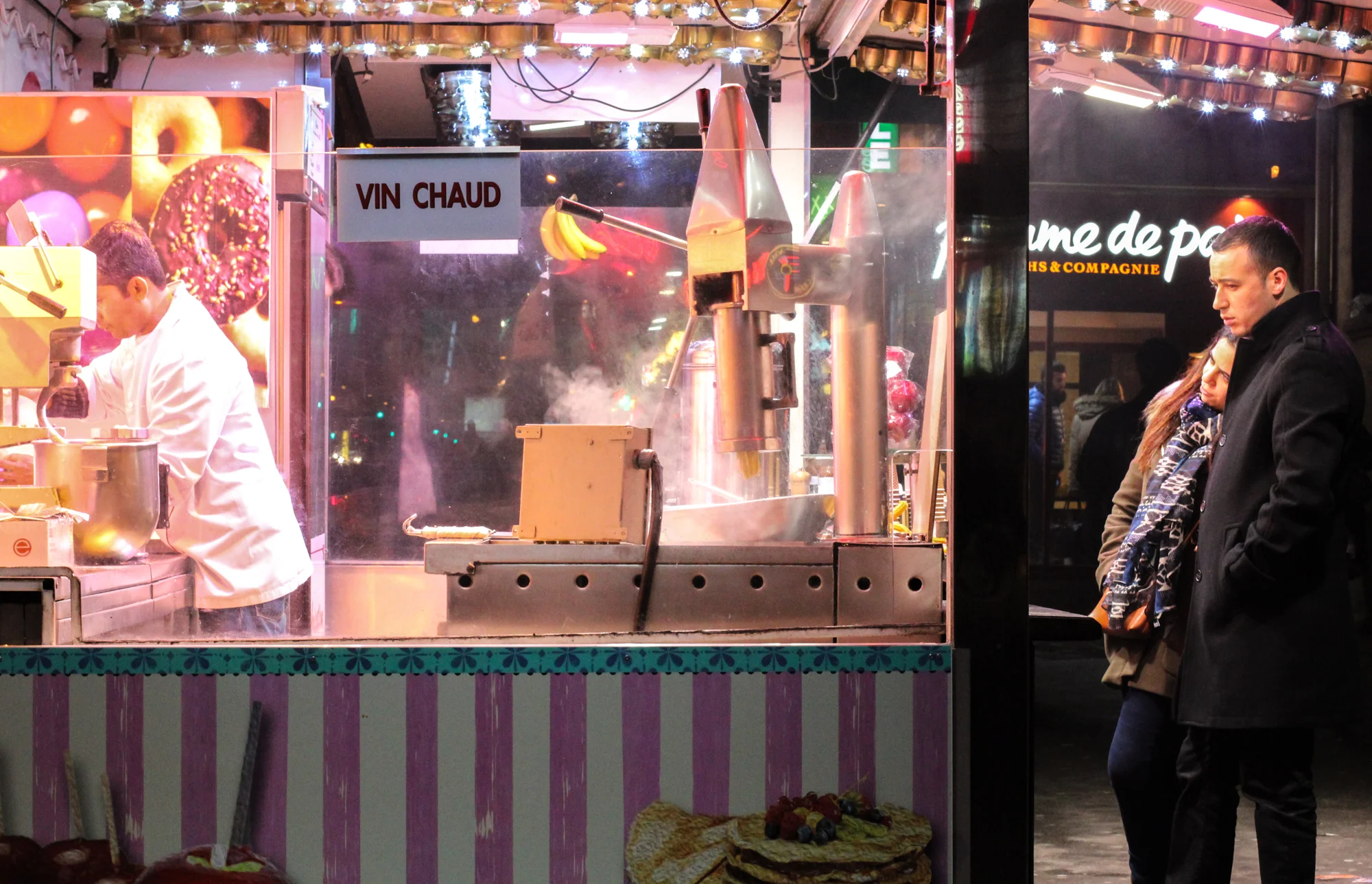 A street food stand selling hot mulled wine, with a man and a woman standing nearby. The stand has a sign that says 'VIN CHAUD,' and the vendor is preparing hot drinks behind the counter. There are colorful decorations and lights in the background.