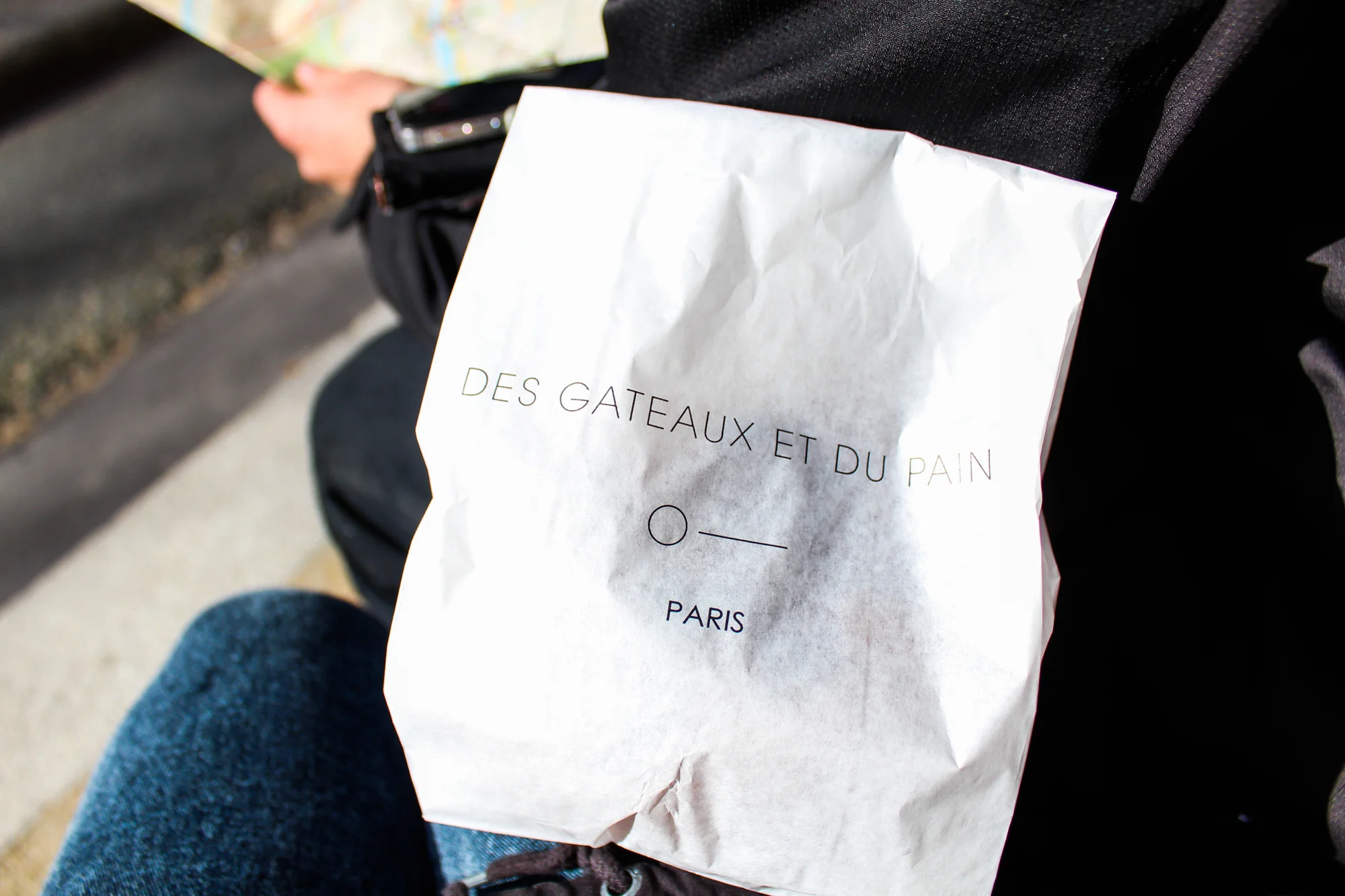 A white paper bag with French text that reads 'Des Gâteaux et du Pain, Paris' resting on a person’s lap outdoors, partially obscuring the person's jeans. The person is wearing a black jacket and is sitting on a bench or ledge.