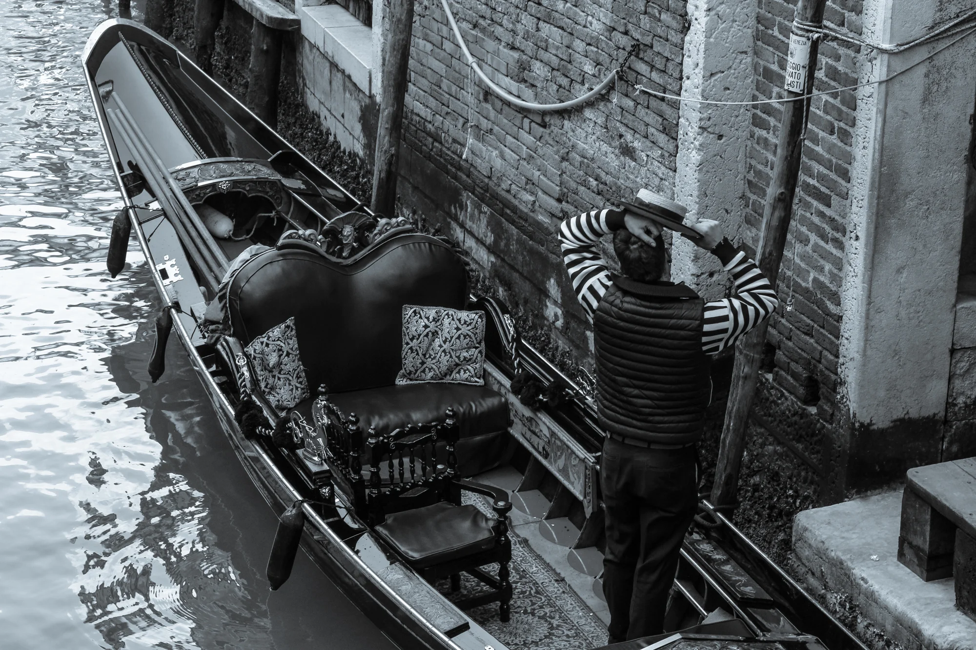 A gondola docked along a canal wall in Venice, Italy, with a man adjusting a hat near the brick wall.
