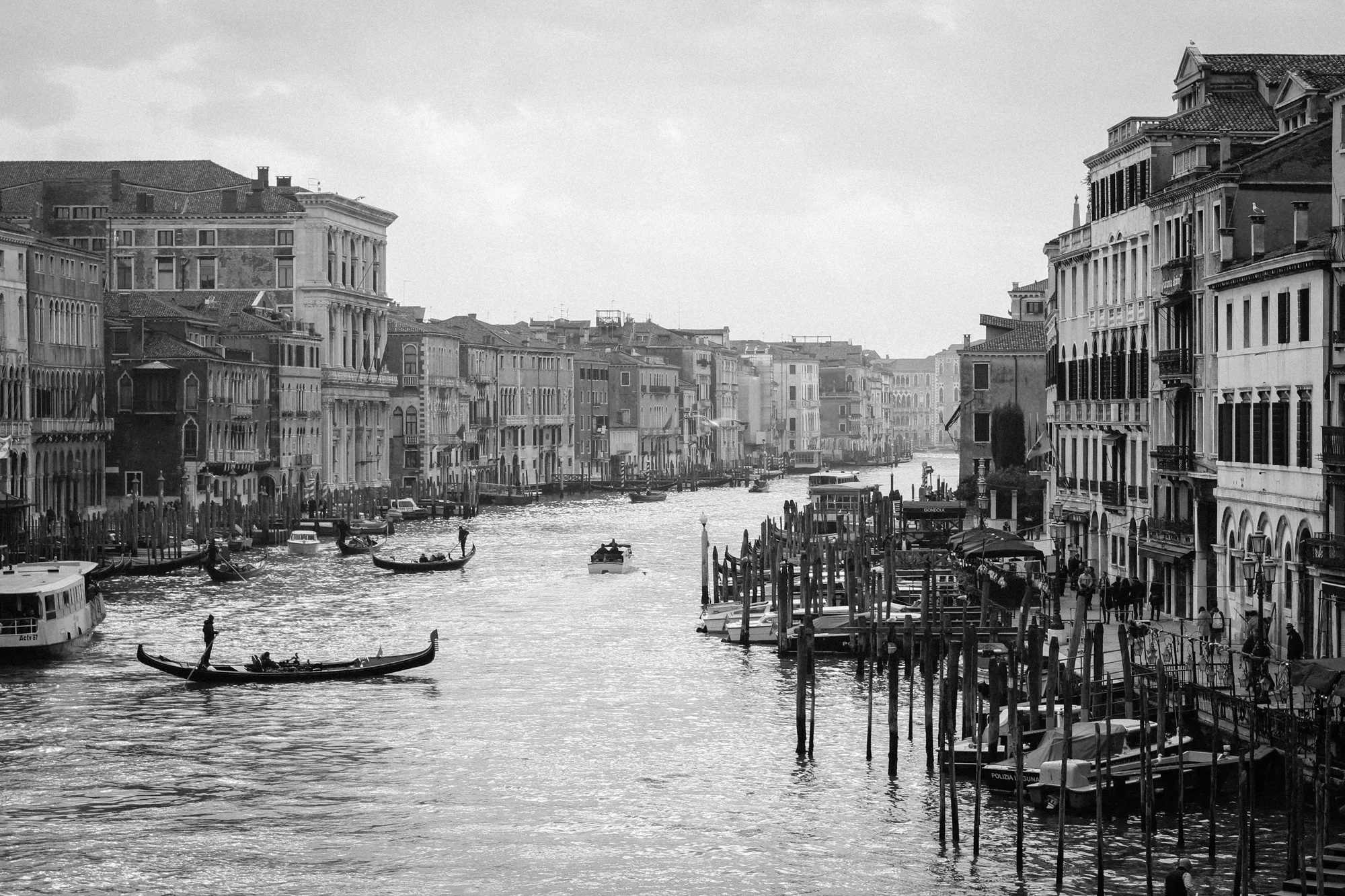 A grayscale photo of a canal with gondolas and boats, historic buildings lining the water, and a cloudy sky.