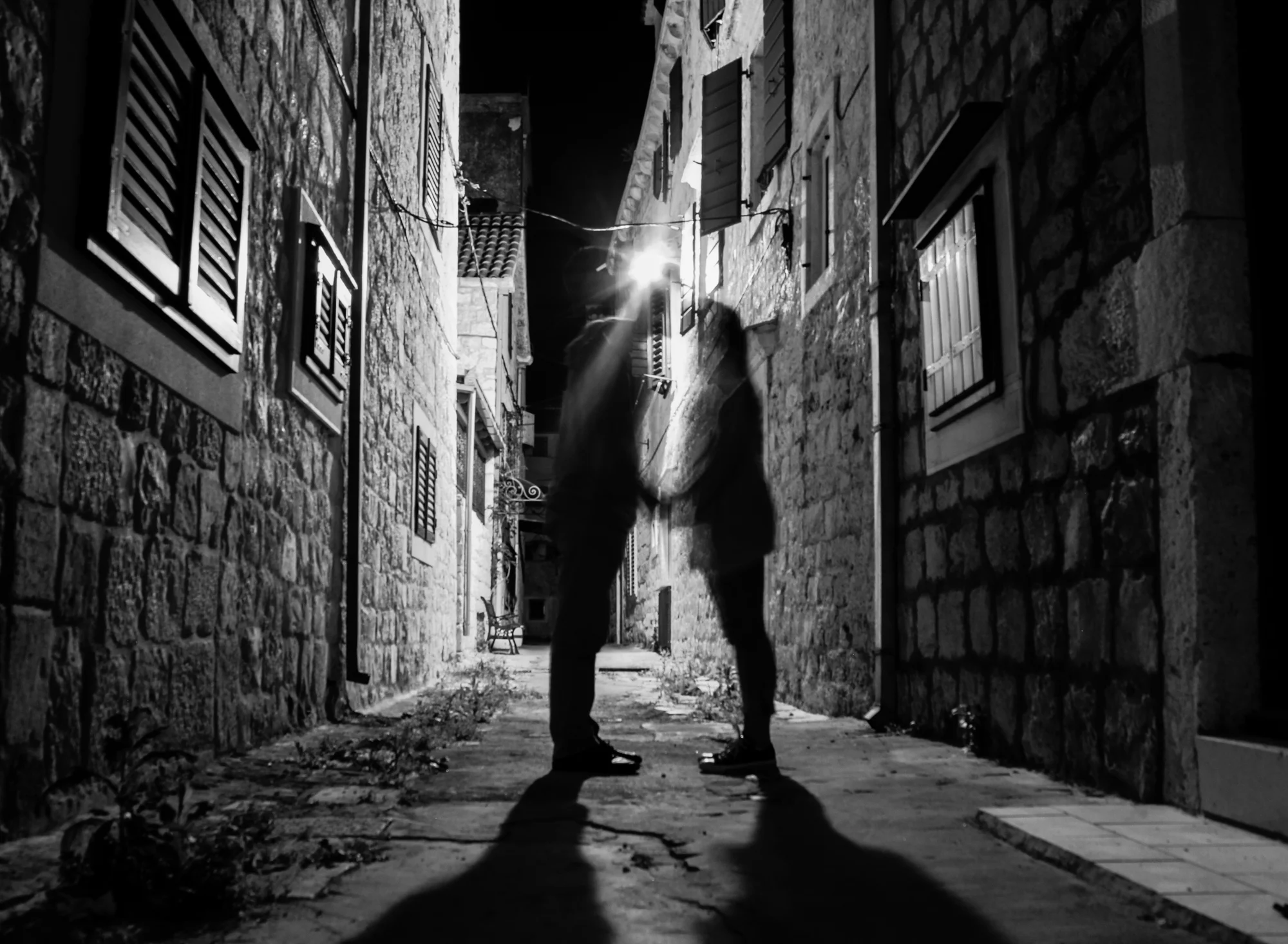 Silhouettes of two people kissing in a narrow alley at night, illuminated by a streetlamp, with brick and stone buildings on either side.