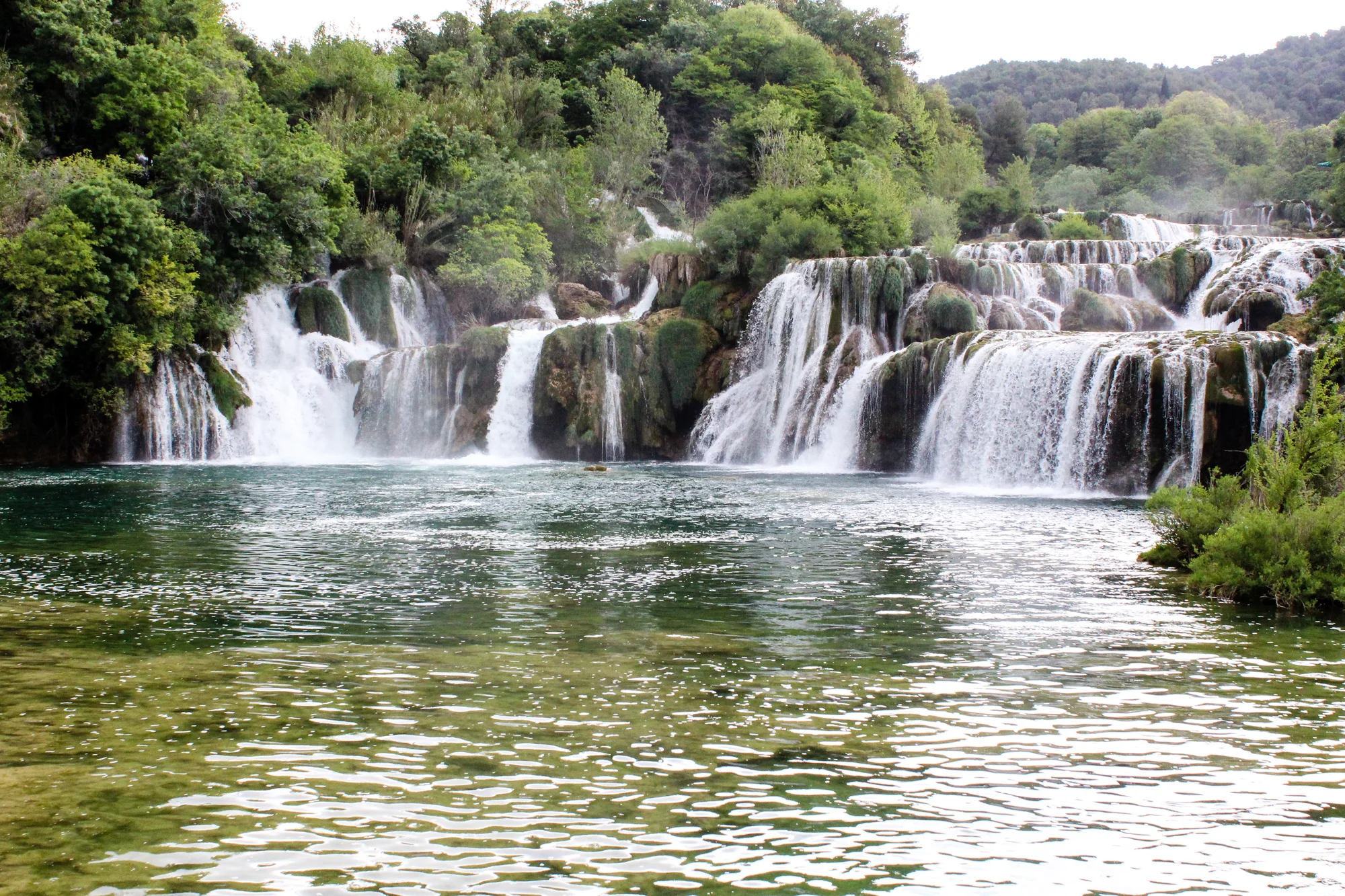Waterfalls flowing over rocks into a calm river surrounded by green trees and hills.