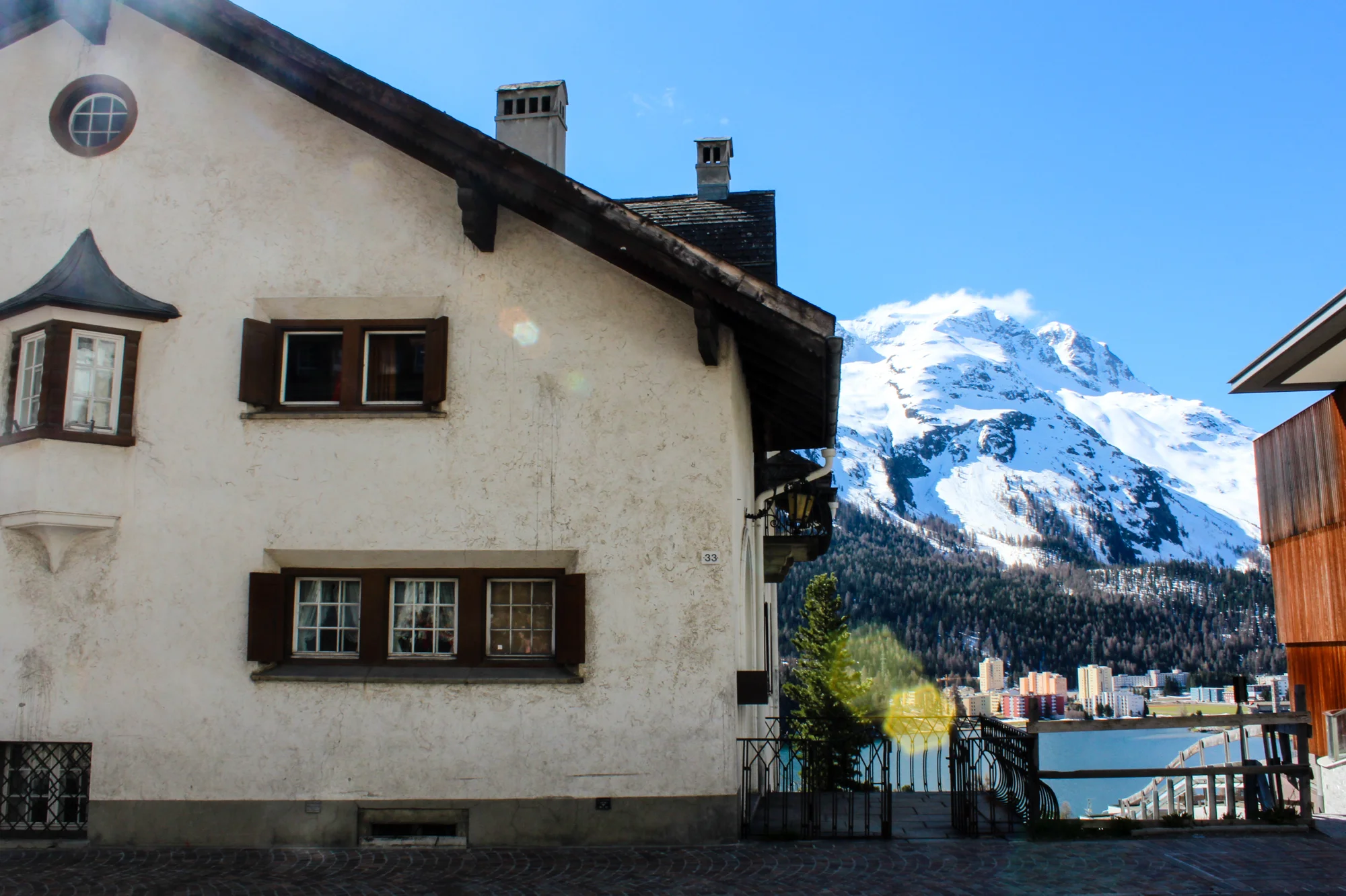 A white building with brown window shutters in front of snow-capped mountain and blue sky.