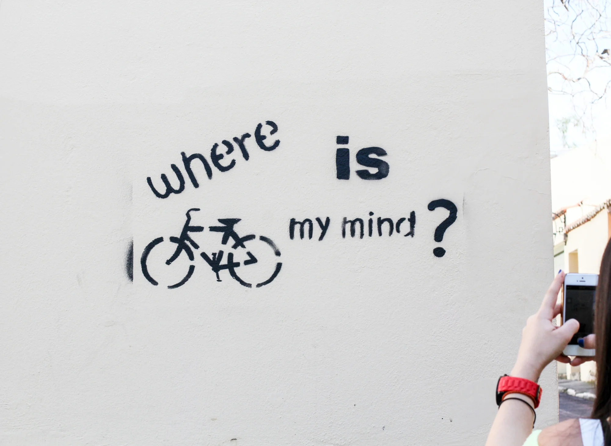 Street art graffiti with text 'where is my mind?' and a bicycle drawn below the text on a white wall, with a woman taking a photo of it.