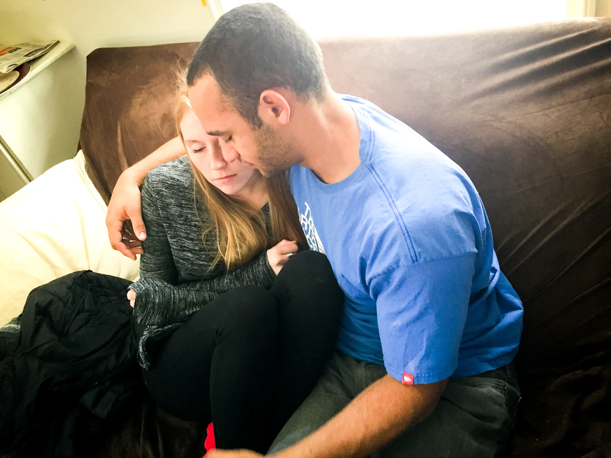 A young man and woman sitting closely on a couch, comforting each other with their eyes closed and arms around each other.