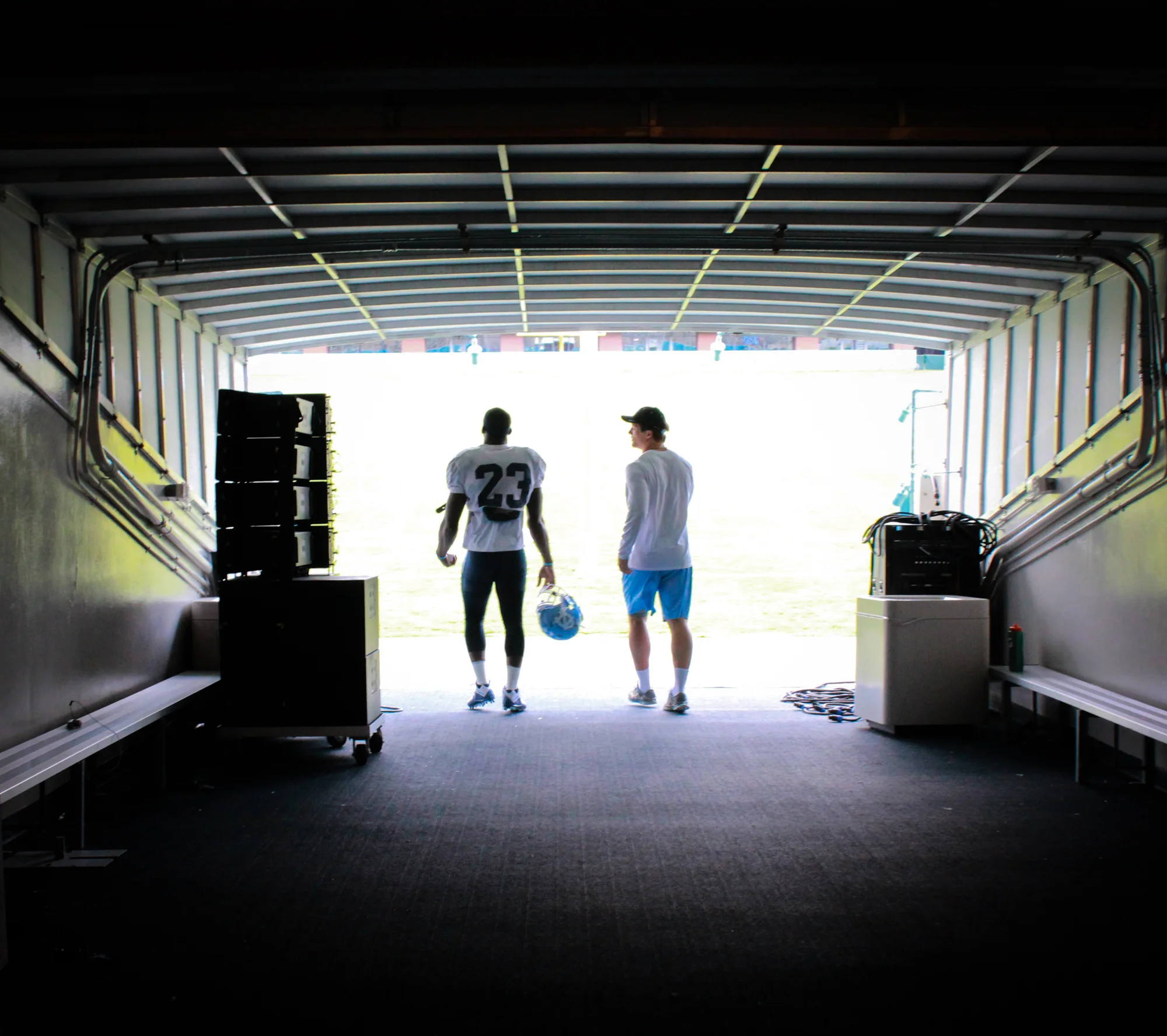 Two football players, one wearing a jersey with the number 23, walking out of a tunnel onto a field, with a coach or teammate walking alongside them.