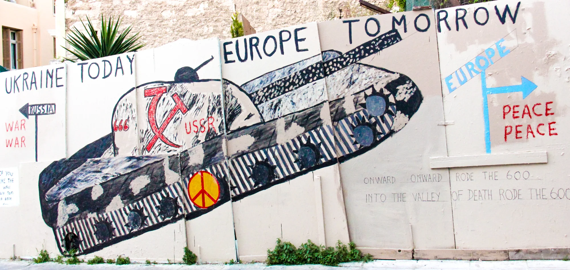 Protest mural on a wall showing a tank with Soviet symbols, with anti-war messages. Text includes "Ukraine Today" with an arrow pointing left to "Russia" and "War War," and "Europe Tomorrow" with an arrow pointing right towards "Peace Peace." The mur