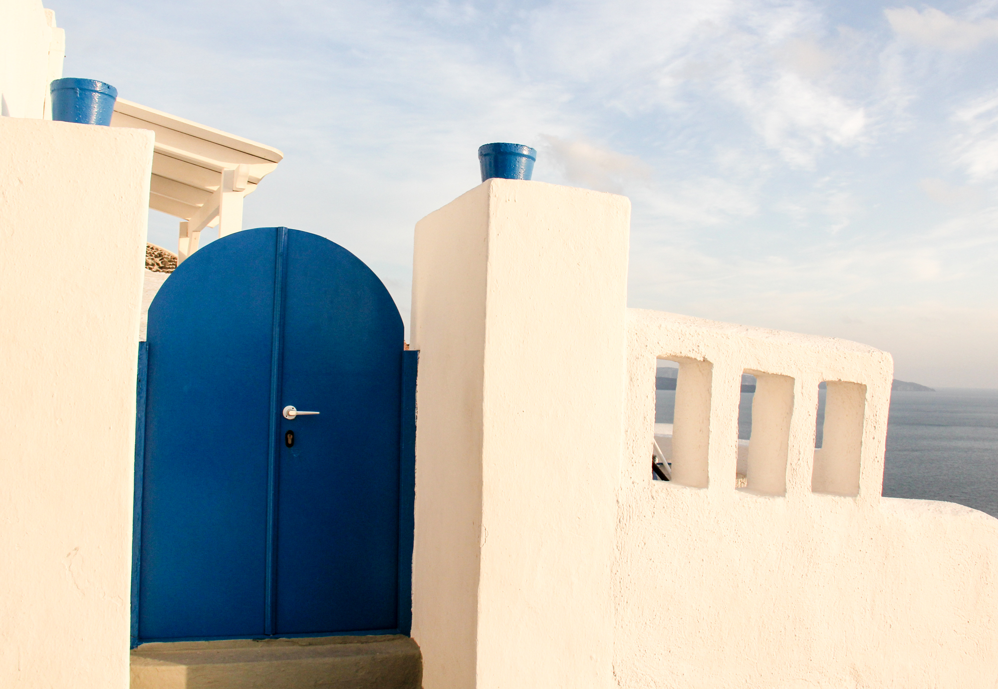 Blue arched door with a silver handle set in a white stucco wall, overlooking a body of water. The wall features three rectangular openings, and there is a blue pot on top of a white structure in the background. The sky is partly cloudy.