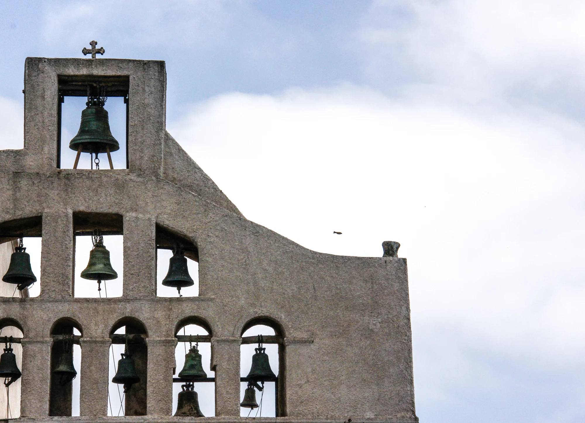 Multiple church bells on a stone wall with a cross at the top, set against a cloudy sky with a small bird flying nearby.