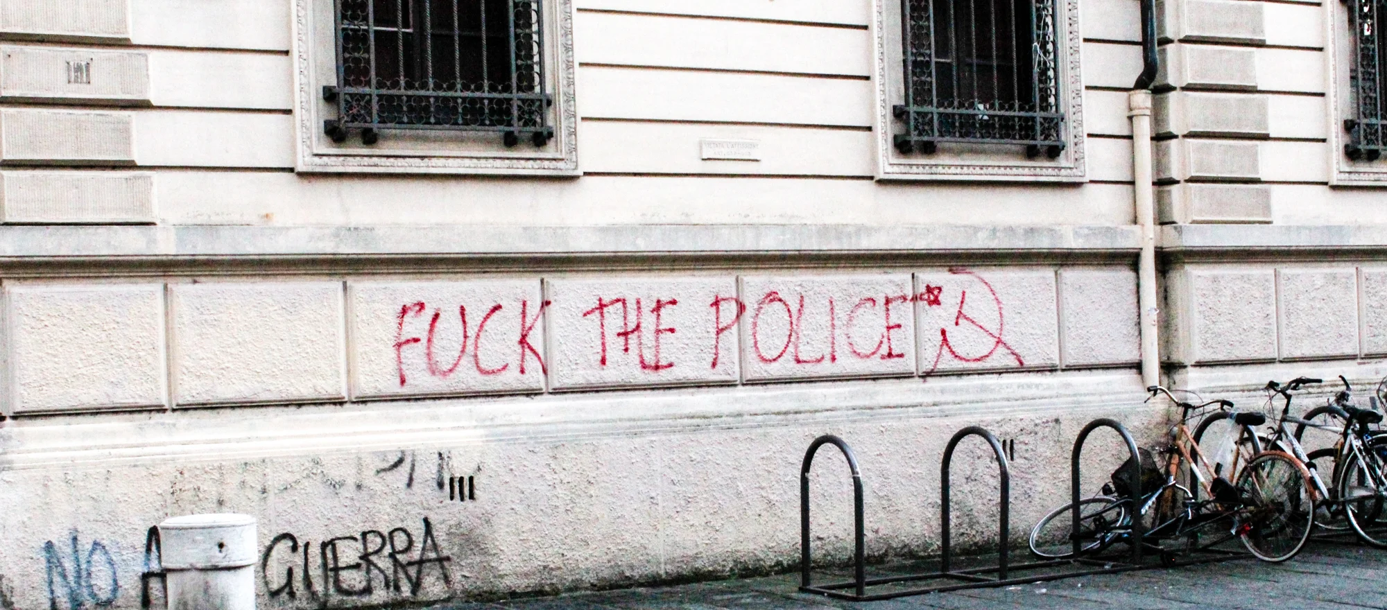 Graffiti on a building wall that reads 'FUCK THE POLICE' in red spray paint, next to bicycles and additional graffiti that says 'NO GERRA' in black paint.