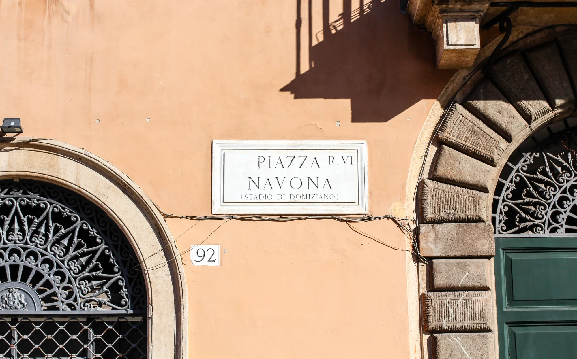 Street sign and architectural details on a building in Pisa, Italy. The sign reads "PIAZZA NAVONA" with a smaller line indicating "STADIO DI DOMIZIANO." The building features ornate iron window grilles and stone bricks around the door. The location i