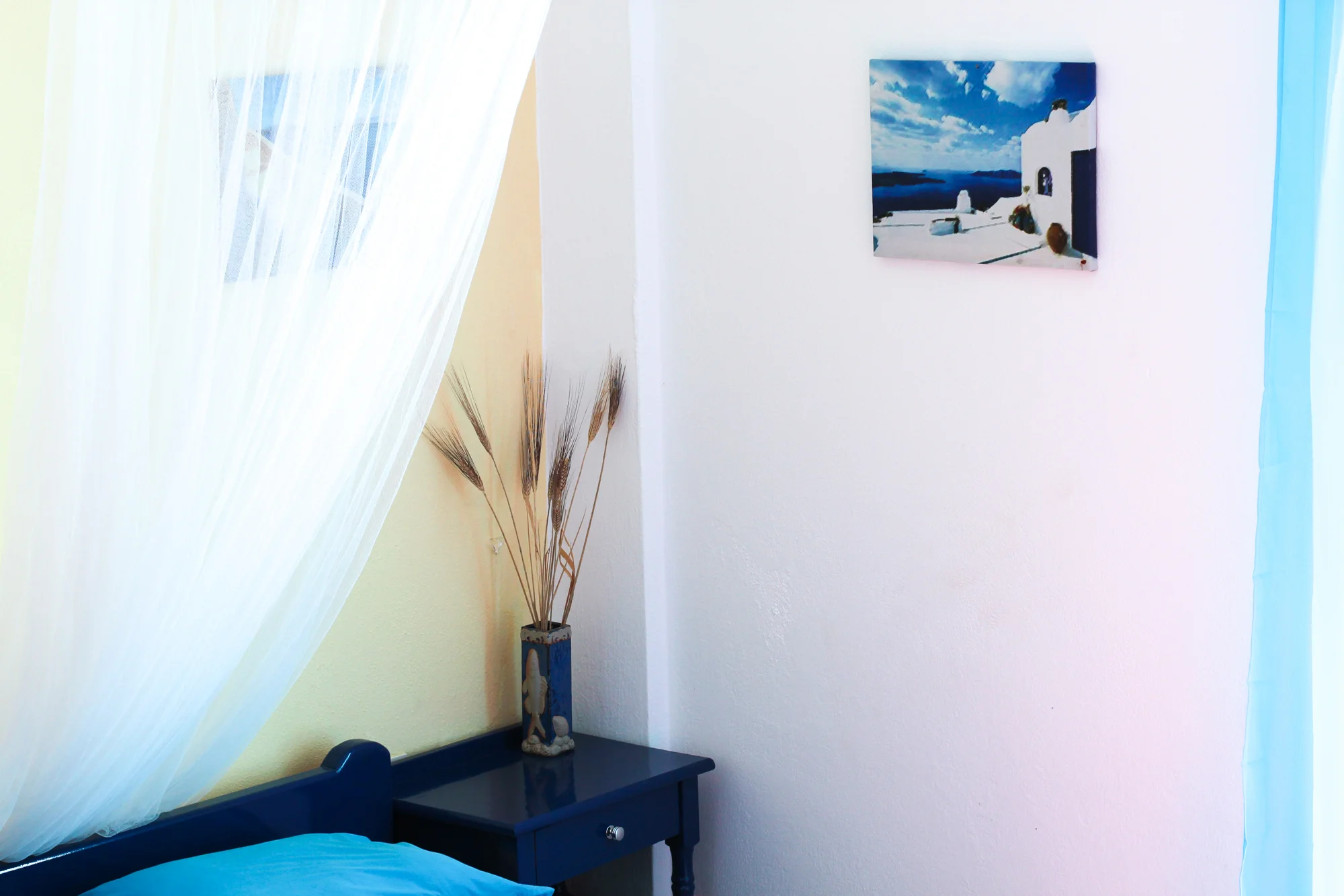 A corner of a bedroom with a blue bed, white sheer curtain, a small dark blue nightstand with a decorative vase with dried wheat stalks, a framed landscape painting on the white wall, and a window with a light blue curtain.